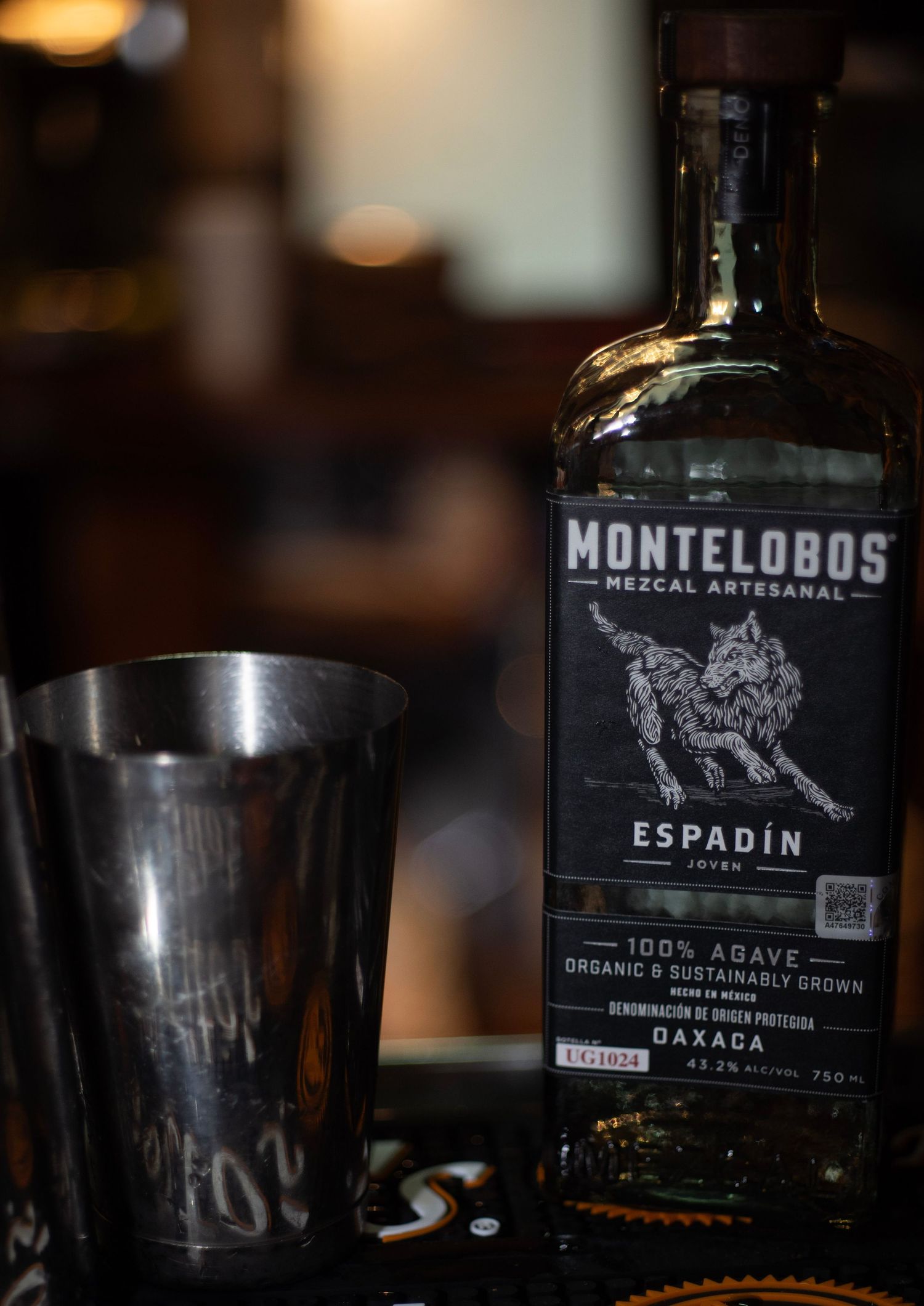Bottle of Montelobos Espadin Mezcal Artesanal beside a silver cocktail shaker in a dimly lit bar setting.