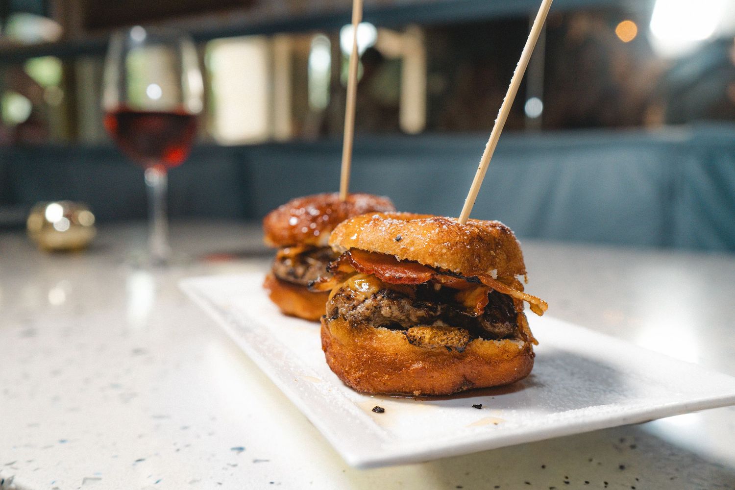 Two mini gourmet sliders on brioche buns served on white board with wine glass in background.