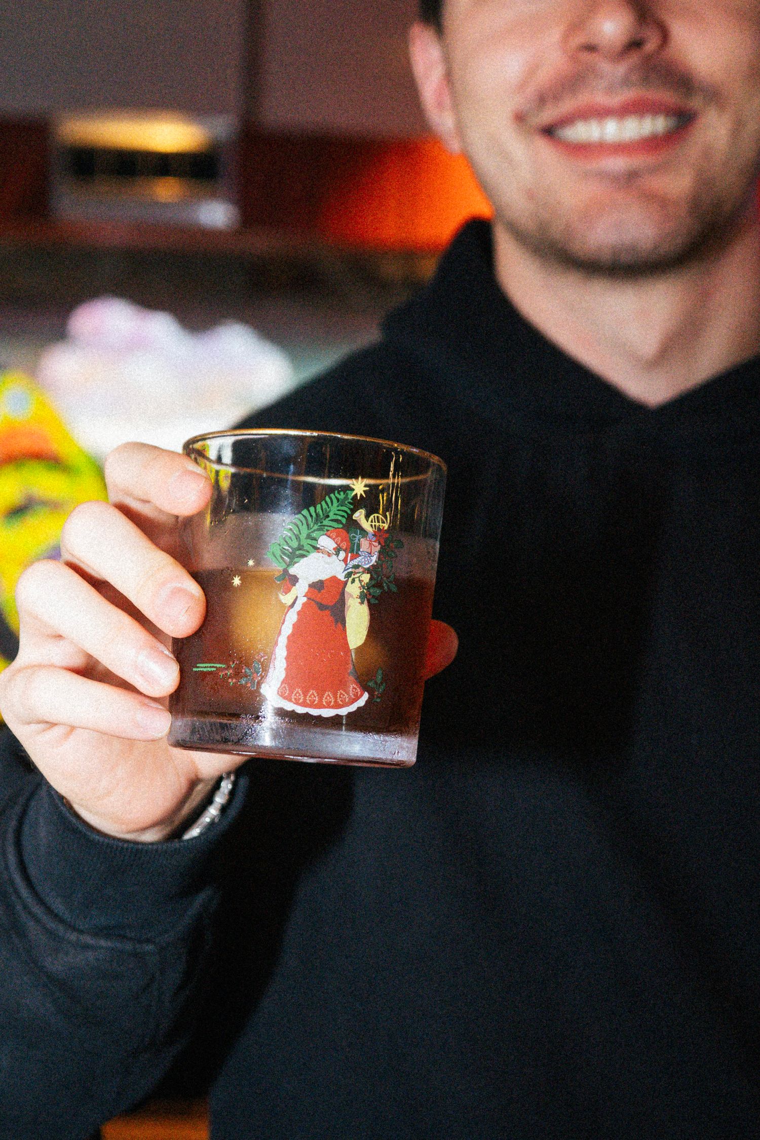 Smiling man in black hoodie holding a vintage Santa Claus Christmas rocks glass with a cocktail inside.