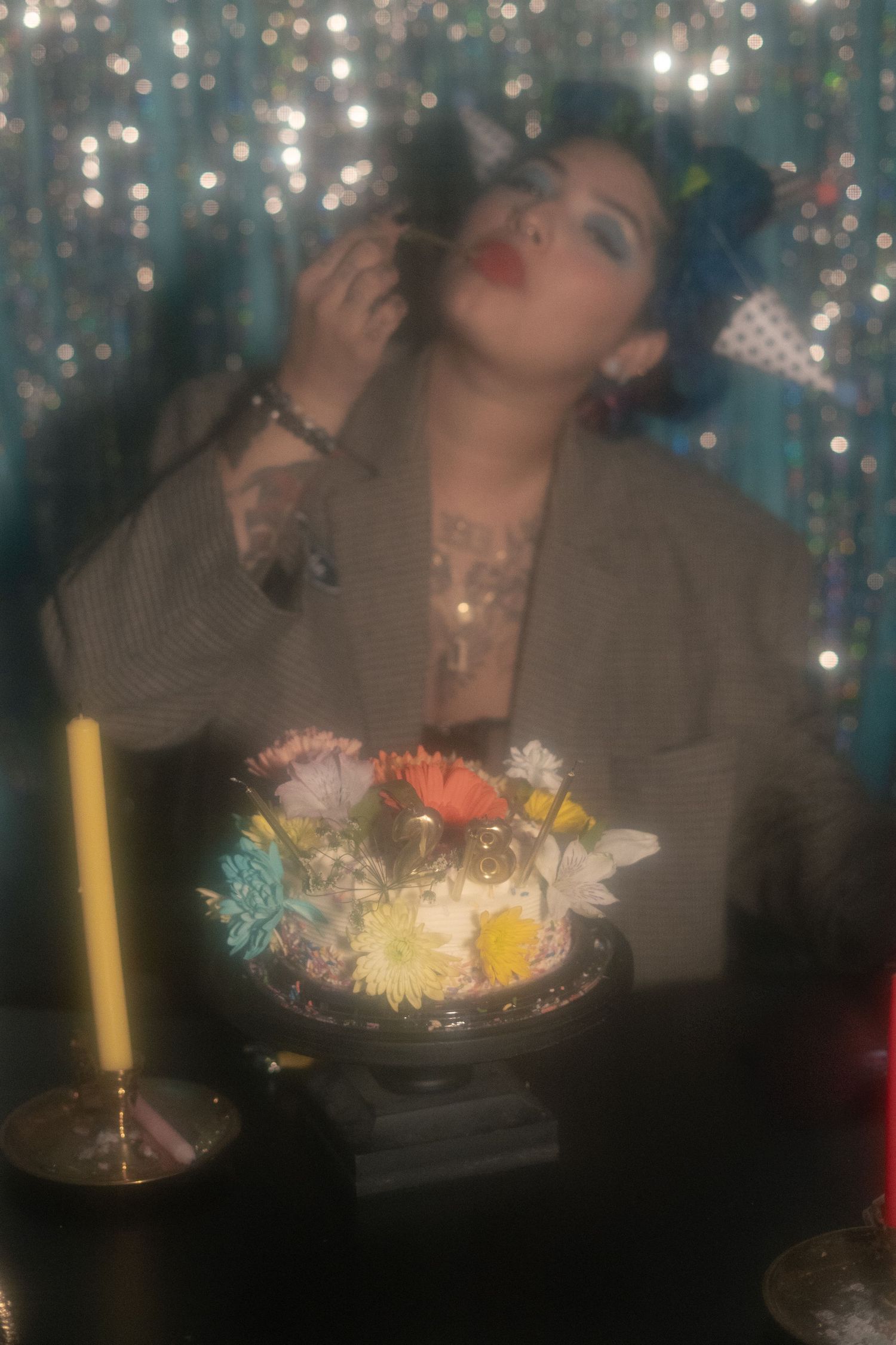 Tattooed woman with blue hair poses with a floral cake and candle against a sparkling teal backdrop.