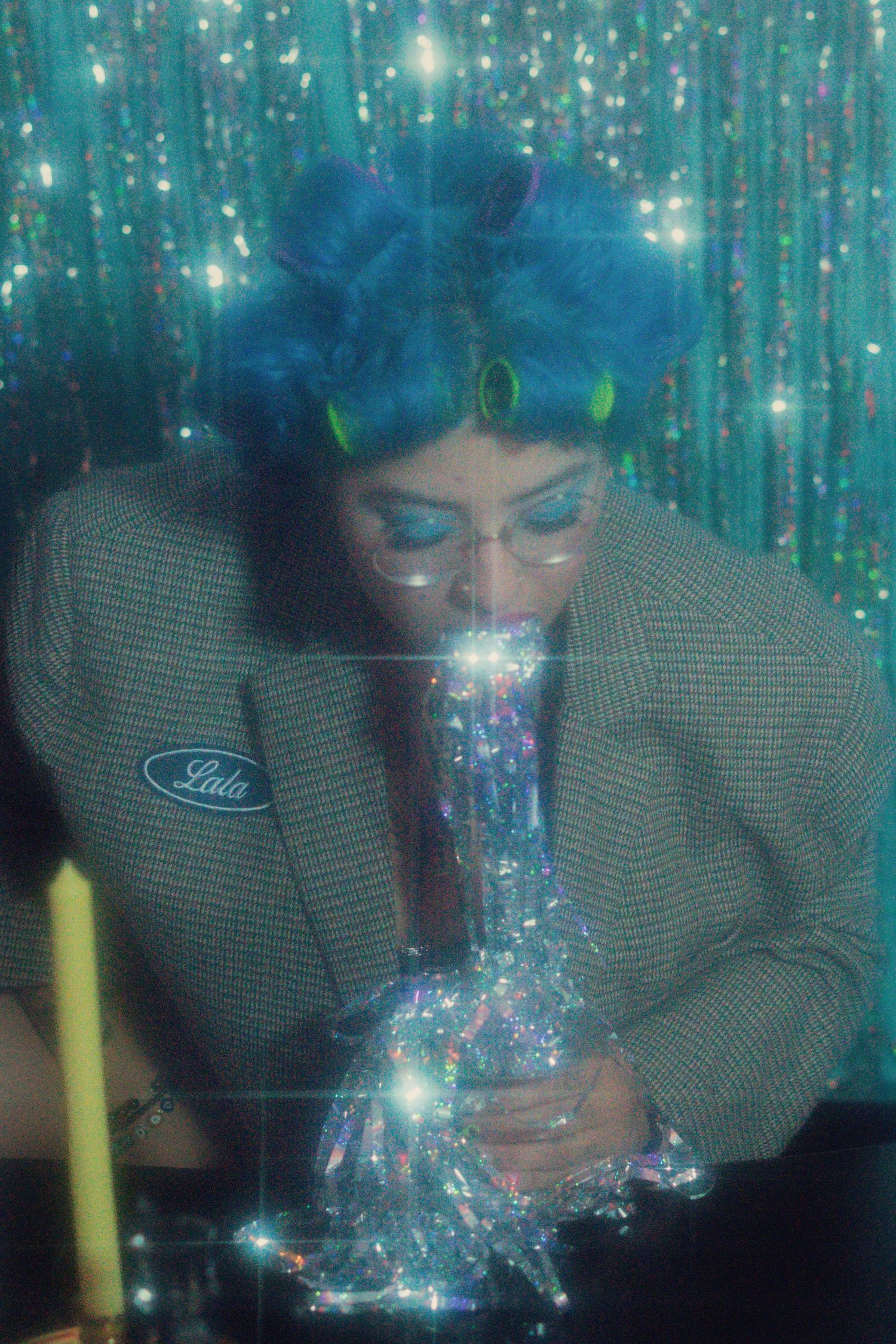 Woman with blue curly hair and green glasses leans over a sparkling object by a candle against a glittery teal backdrop.