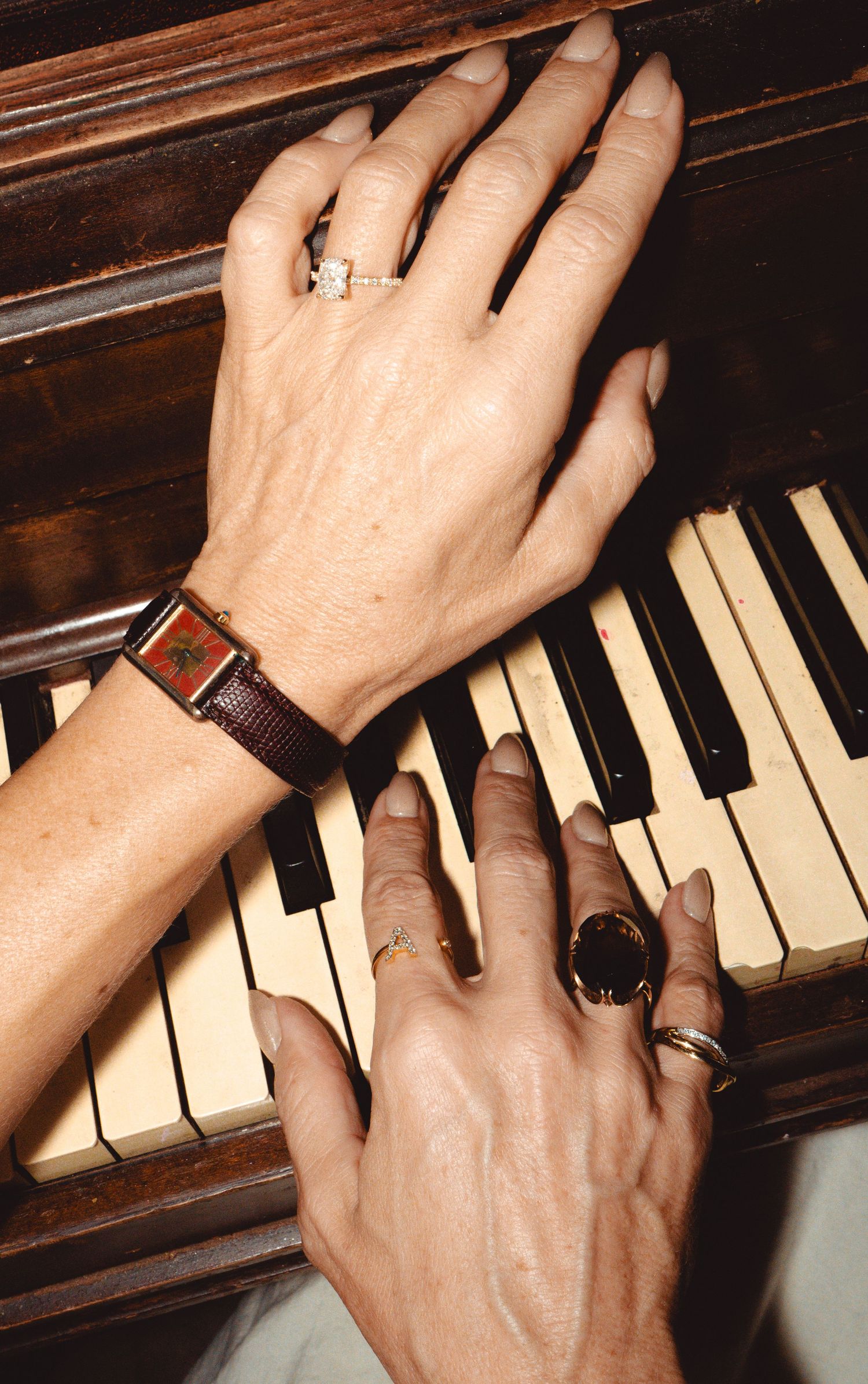 Two hands adorned with rings and a watch playing piano keys on a vintage wooden upright piano.