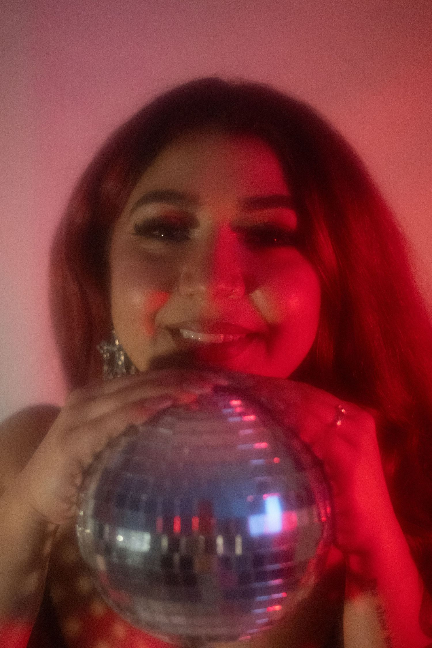 Smiling woman with red hair holding silver disco ball under warm red lighting.