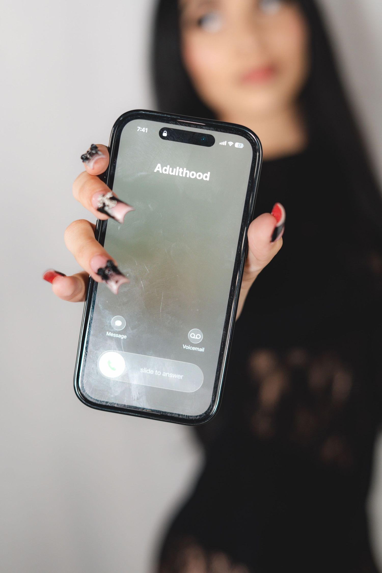 Young woman in black lace dress holding a phone displaying the word Adulthood toward camera.