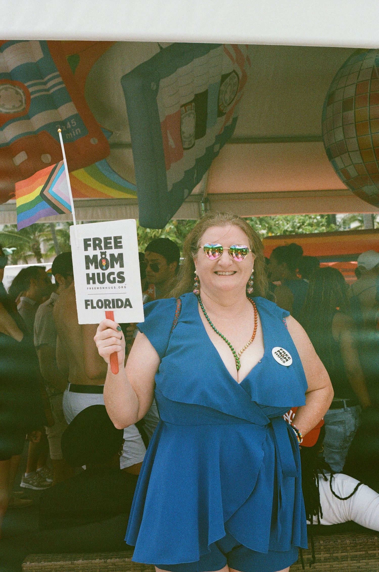 Woman in blue dress holds Free Mom Hugs sign with pride flag at Florida LGBTQ+ Pride event.
