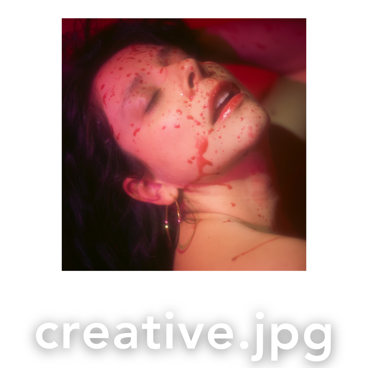 Person with dark hair lying down with red paint splattered across their face and neck in artistic photography.