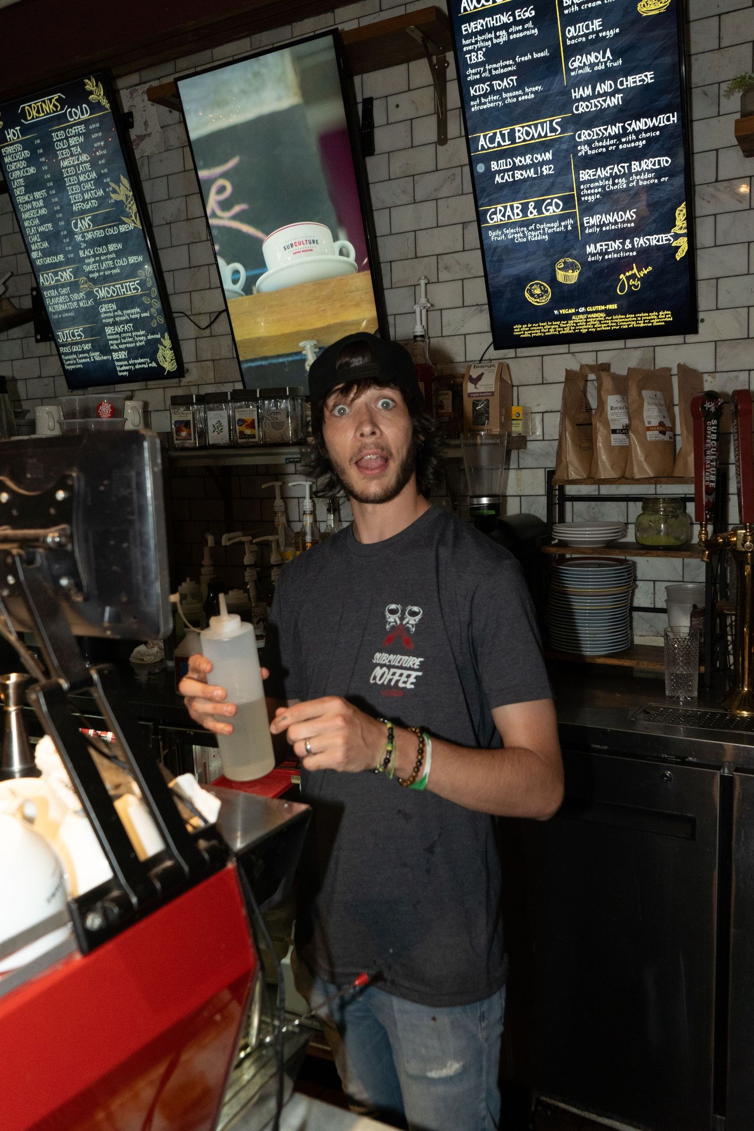A barista in a dark coffee shop t-shirt holds a bottle while standing behind the counter with menu boards above.