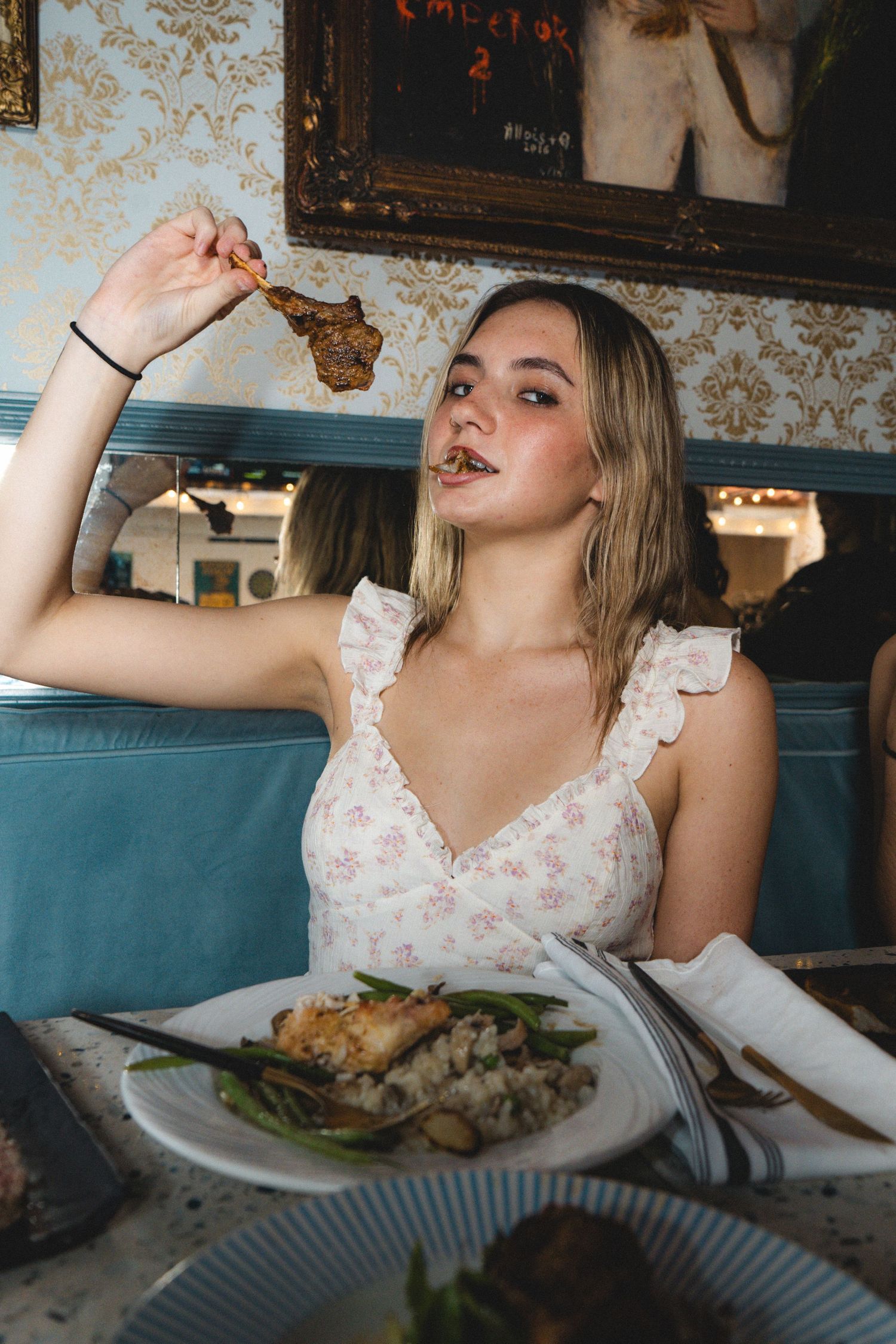 Blonde woman in floral dress eating meat dish at elegant restaurant with blue velvet seating.
