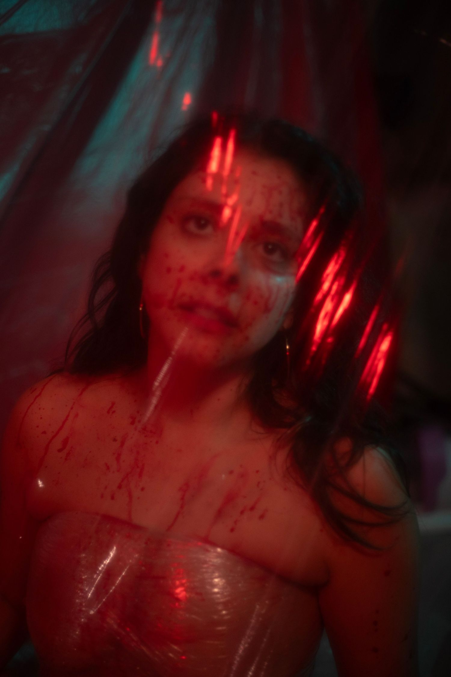 A woman covered in simulated blood poses behind plastic wrap under dramatic red lighting in a horror shoot.