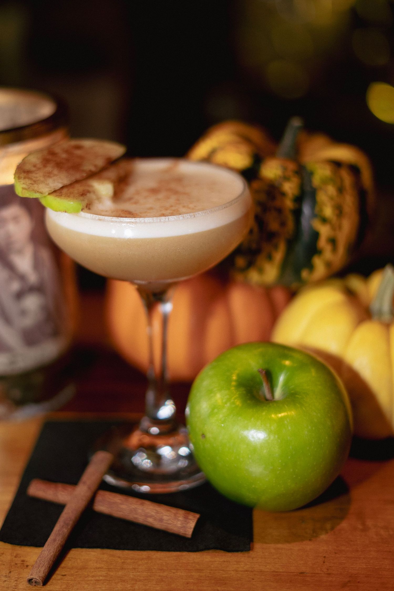 Autumn cocktail in coupe glass with apple garnish and cinnamon sticks beside a green apple and decorative pumpkins.