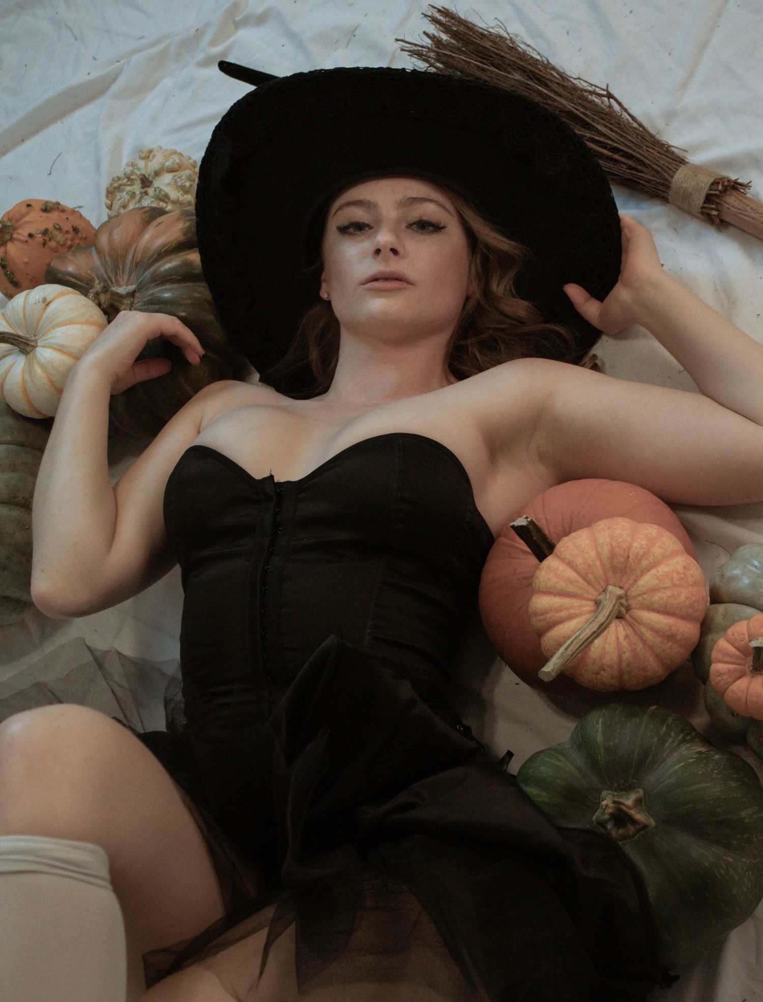 Woman in black strapless dress and witch hat lying among assorted pumpkins in a Halloween-themed photo.