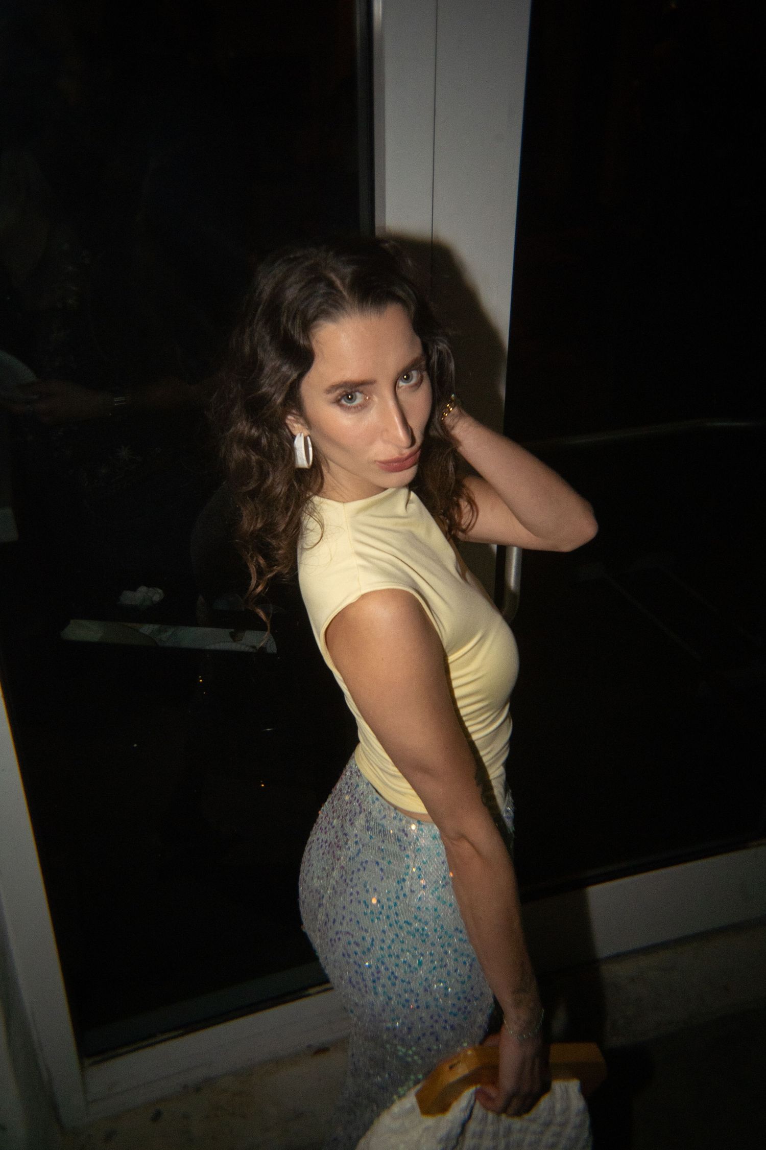 Young woman in yellow top and sequin skirt posing indoors at night, looking over shoulder with curly hair.