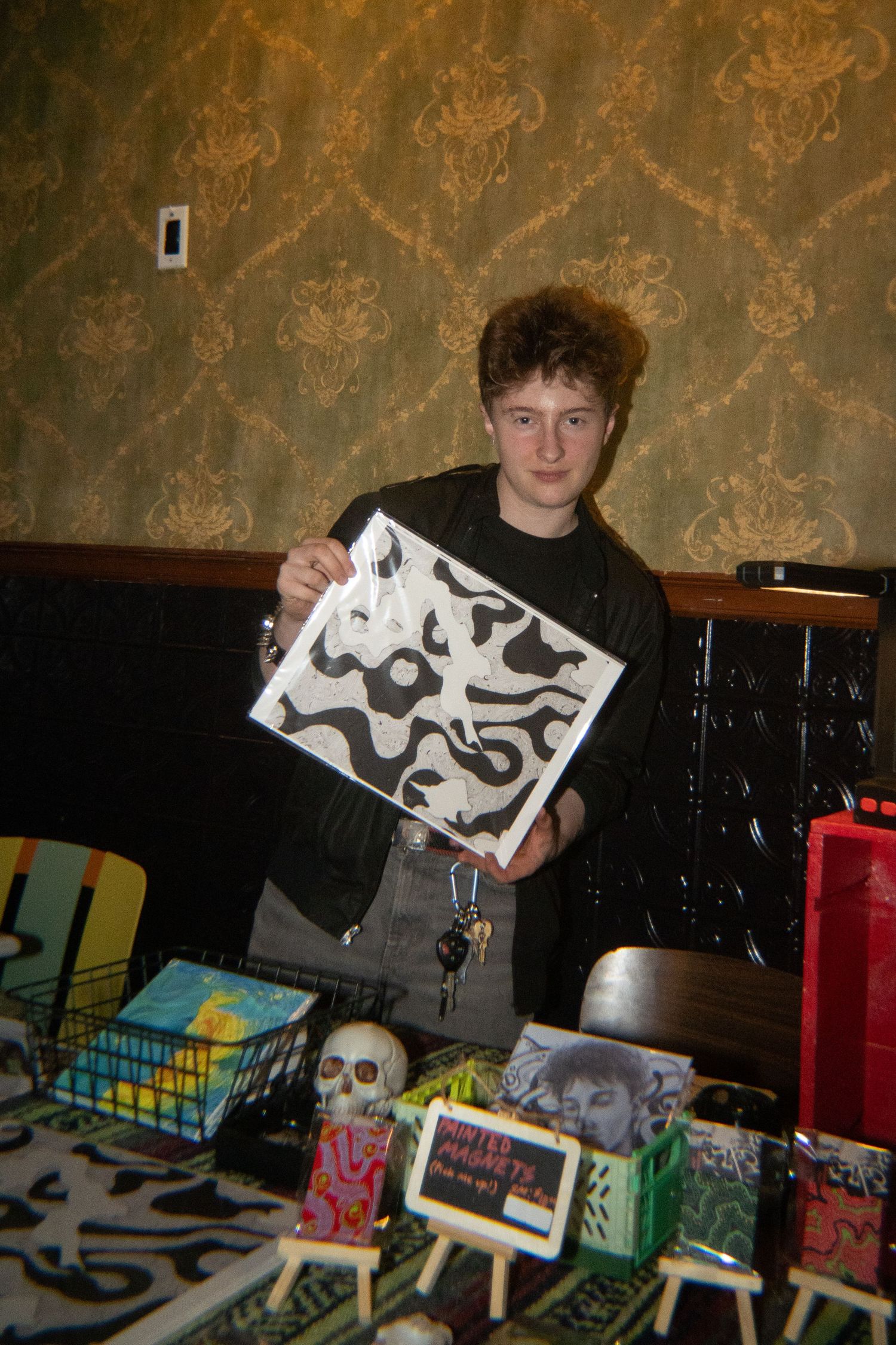 Young artist displays black and white abstract artwork at an indoor art fair table with skulls and colorful mini paintings.