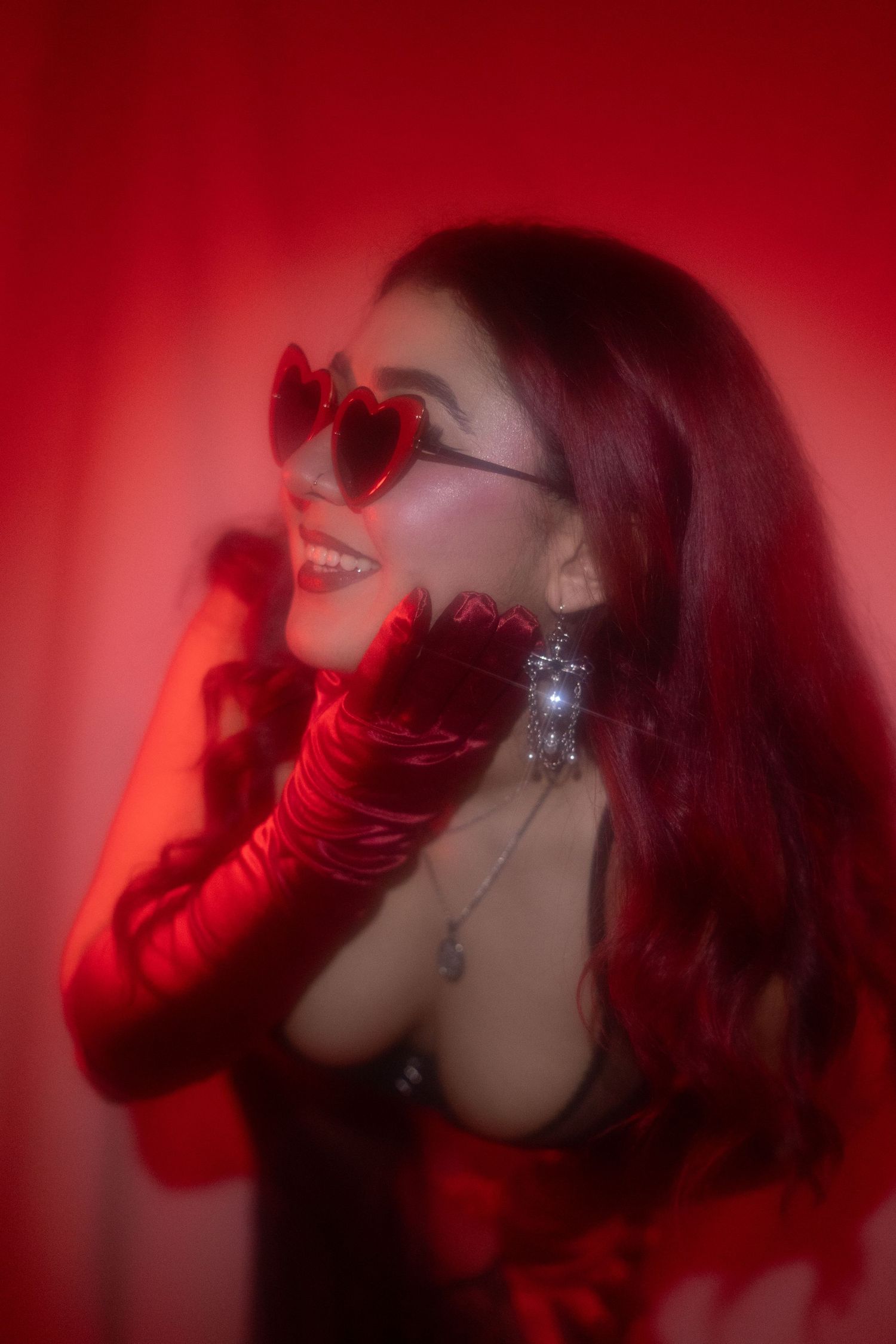 Smiling woman with long red hair wearing heart-shaped sunglasses and red satin gloves in red lighting.