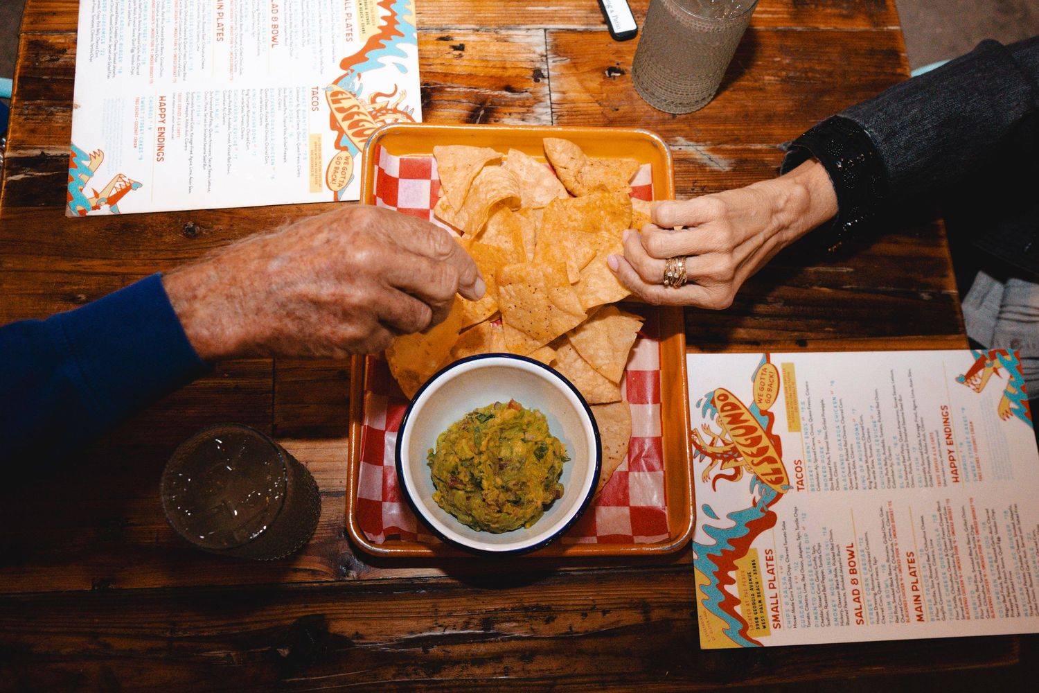 Two people sharing chips and fresh guacamole served on a red checkered tray at a casual Mexican restaurant.