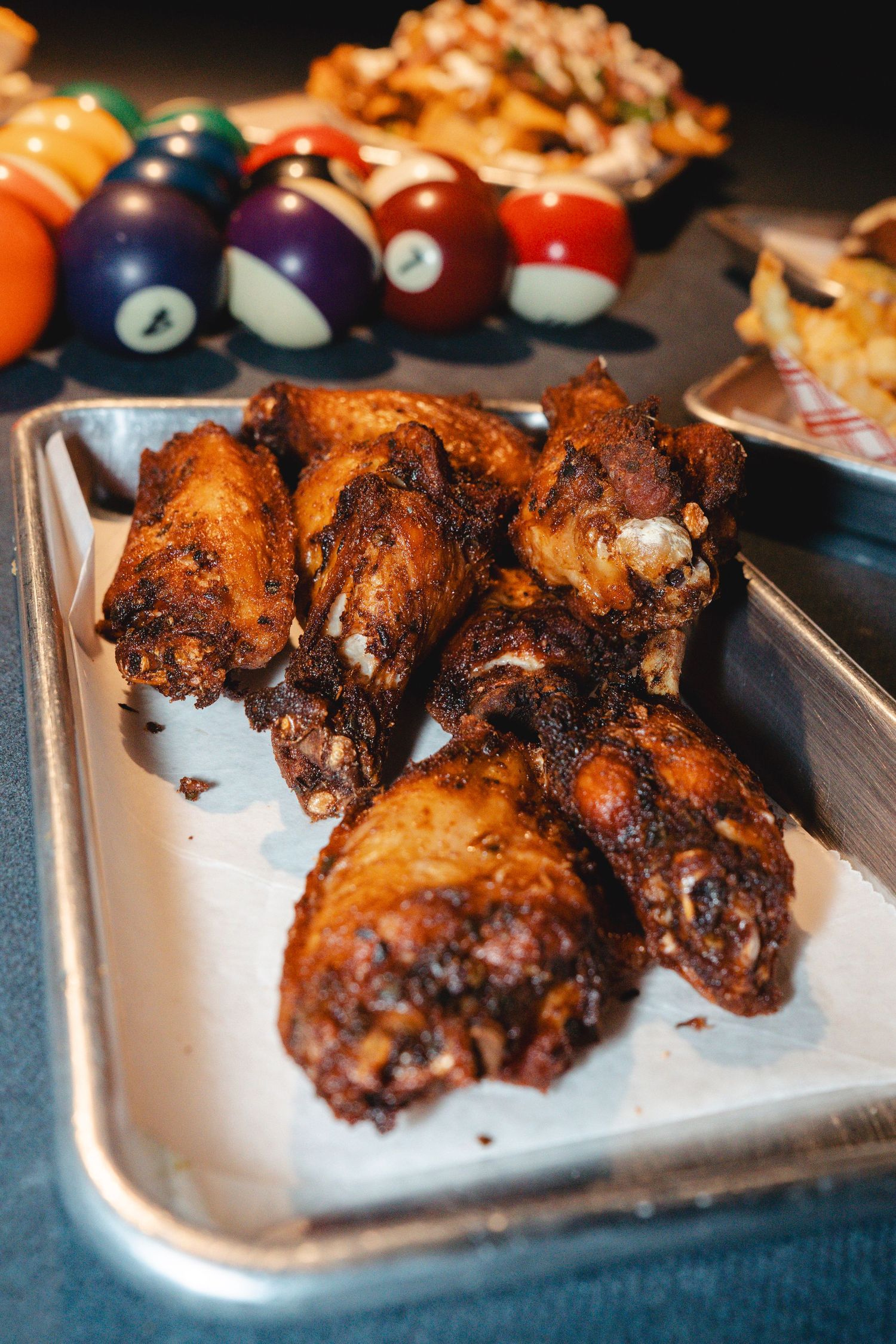 Crispy baked chicken wings on a metal tray beside colorful billiard balls on a pool table.