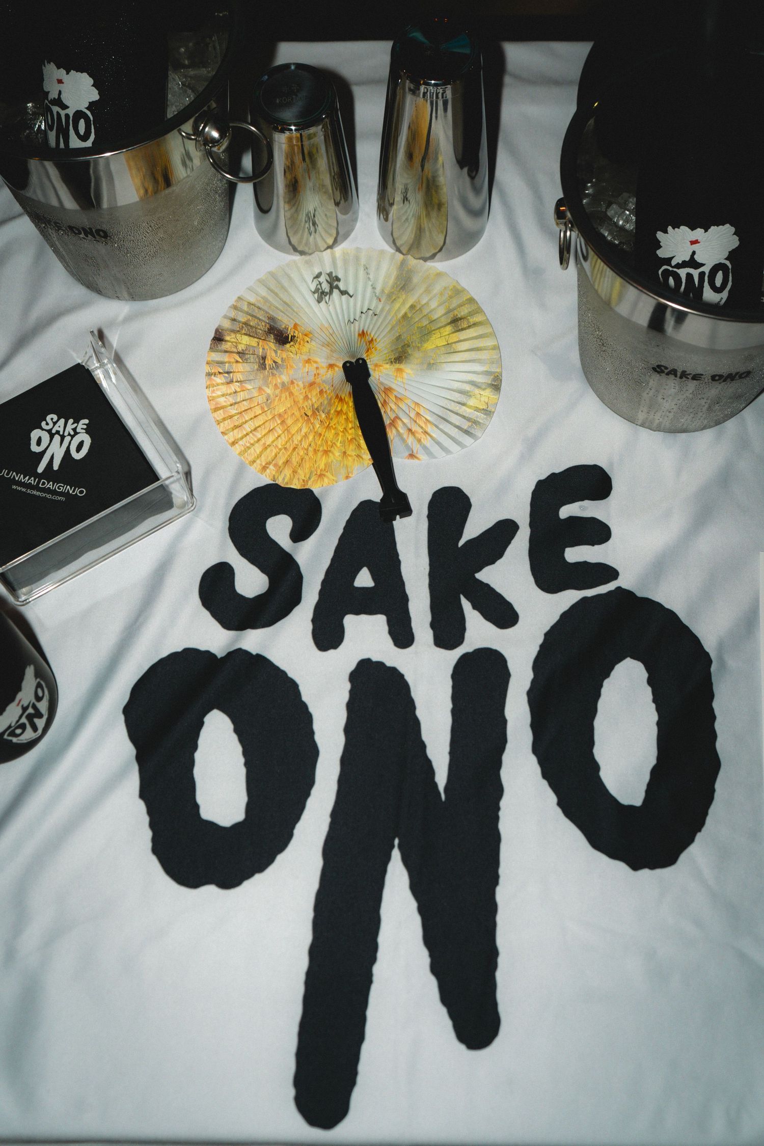 Sake ONO branded merchandise including hats, glasses, fan, and t-shirt displayed on white table.