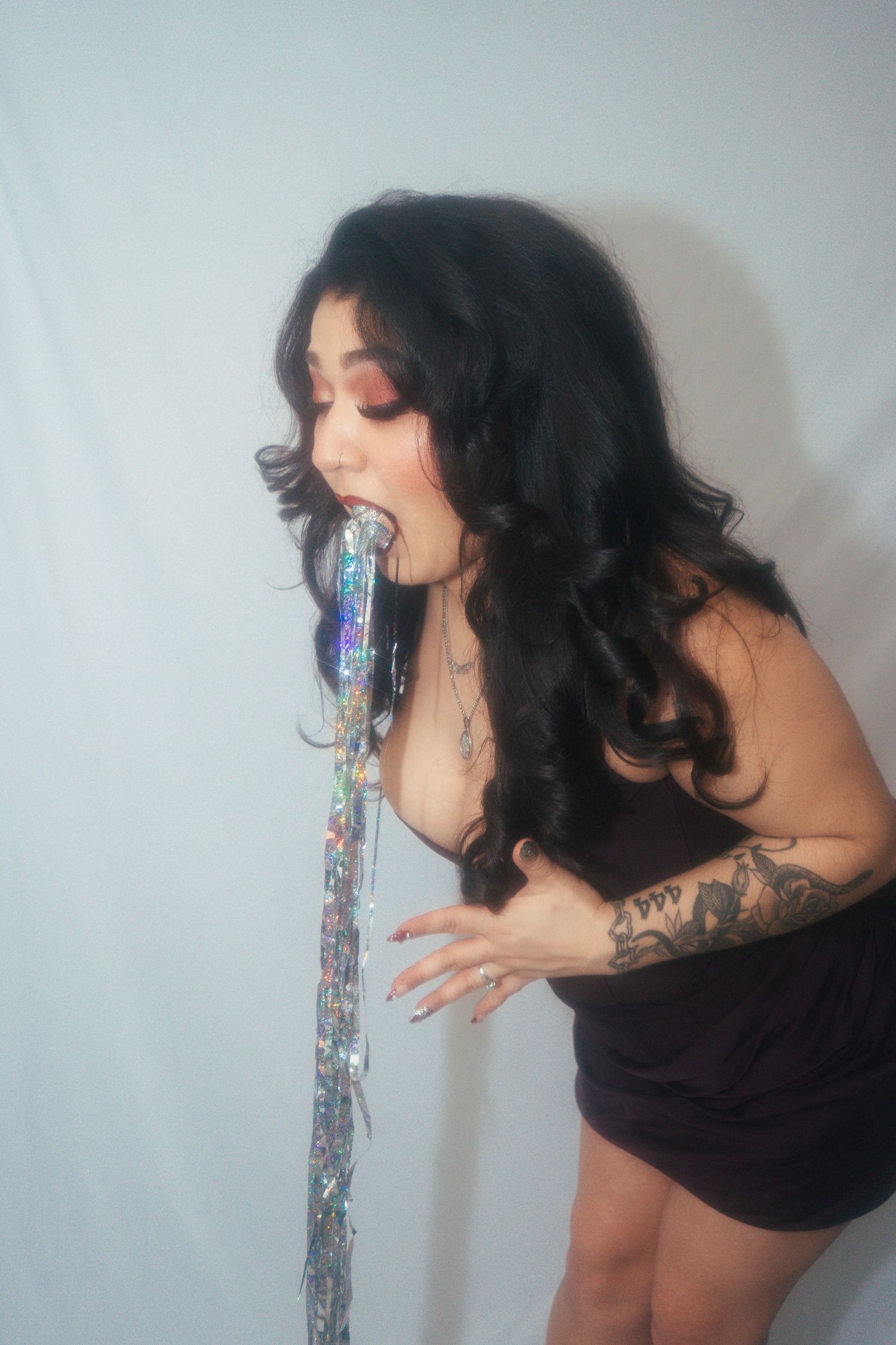 Woman in black mini dress posing with holographic tinsel streamer against white backdrop.