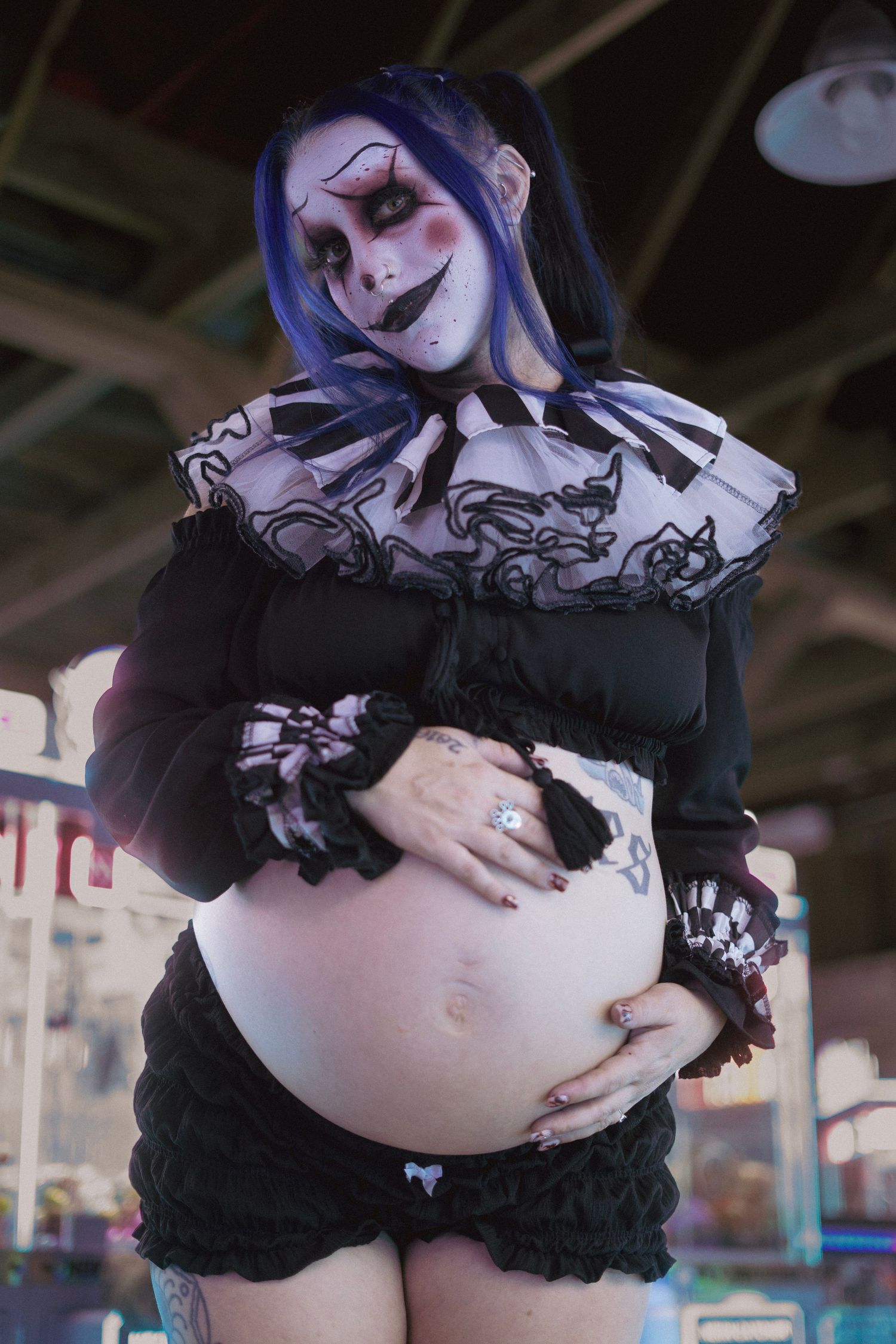 Pregnant person in clown makeup with blue hair cradling baby bump at carnival.