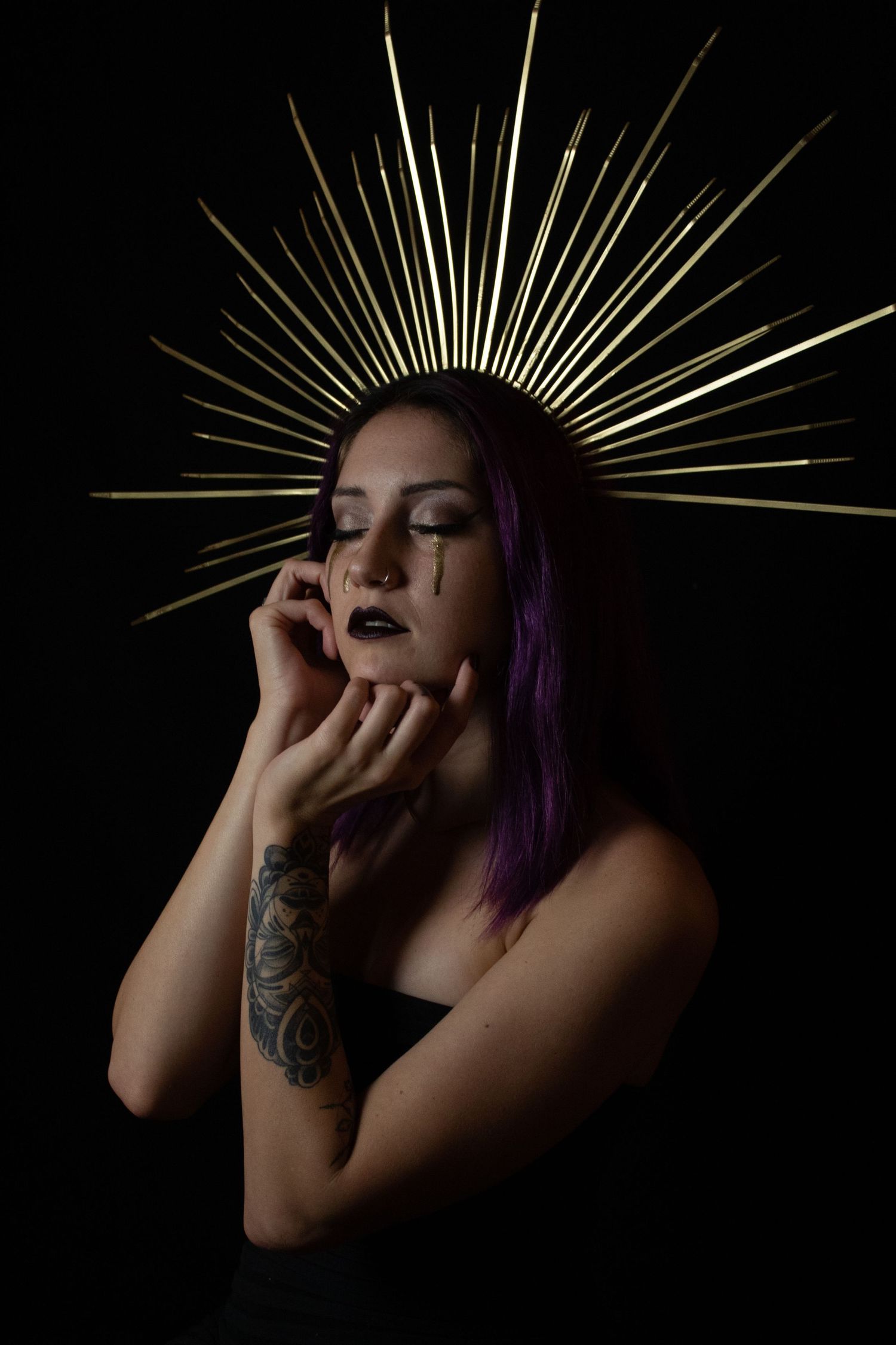 Woman with purple hair wears gold spiked halo crown against black background.