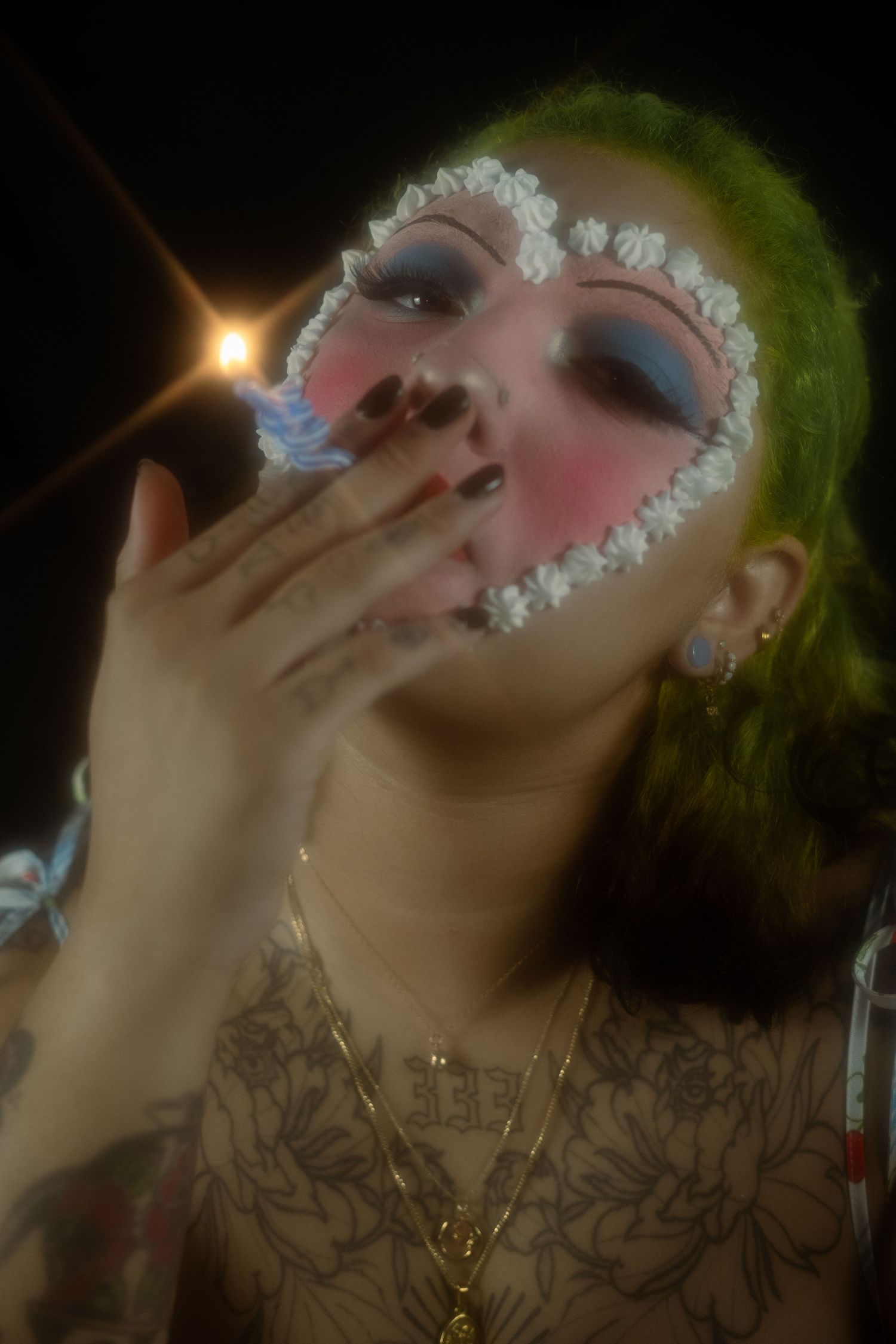 Person with green hair and floral face makeup lighting a cigarette against dark background with chest tattoos visible.