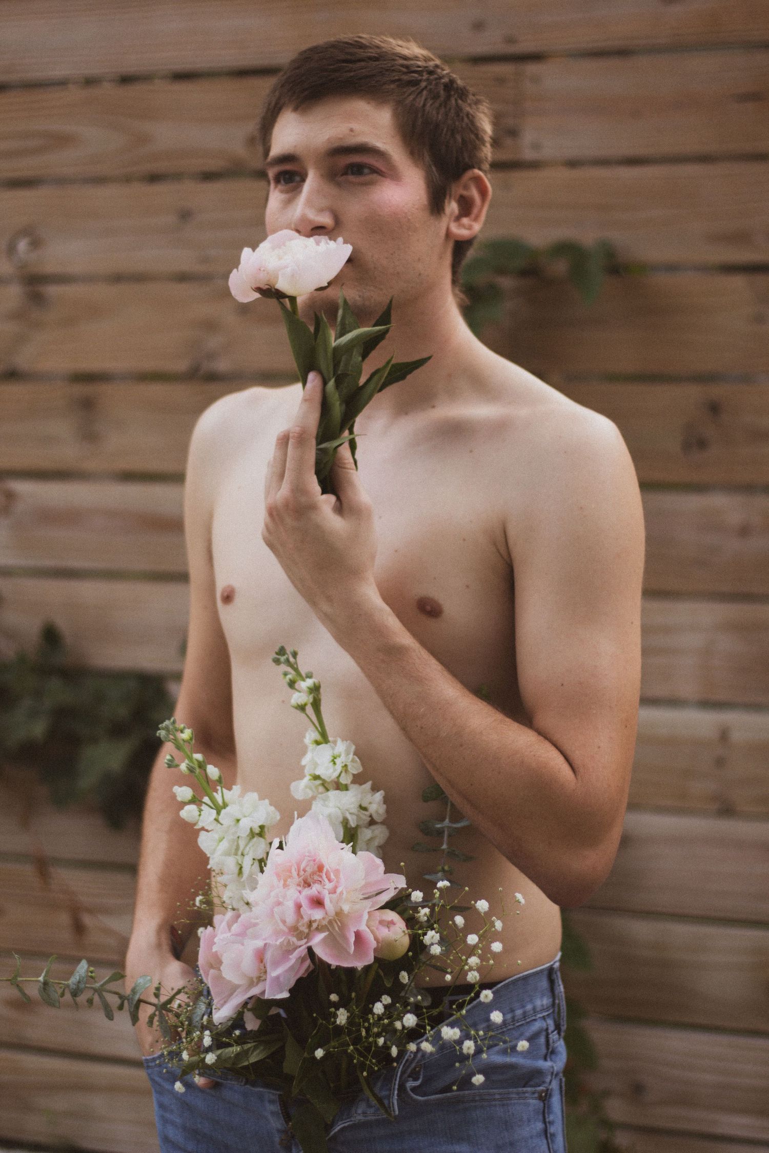 Shirtless young man smelling a white peony while holding a bouquet of pink and white flowers outdoors.