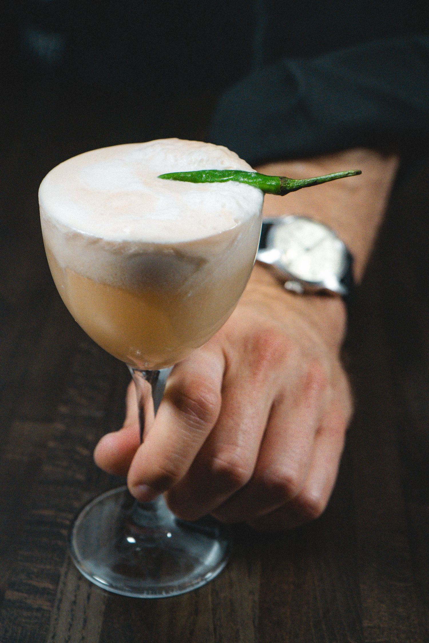 Hand holding elegant cocktail glass with frothy drink garnished with green chili pepper on dark wood surface.