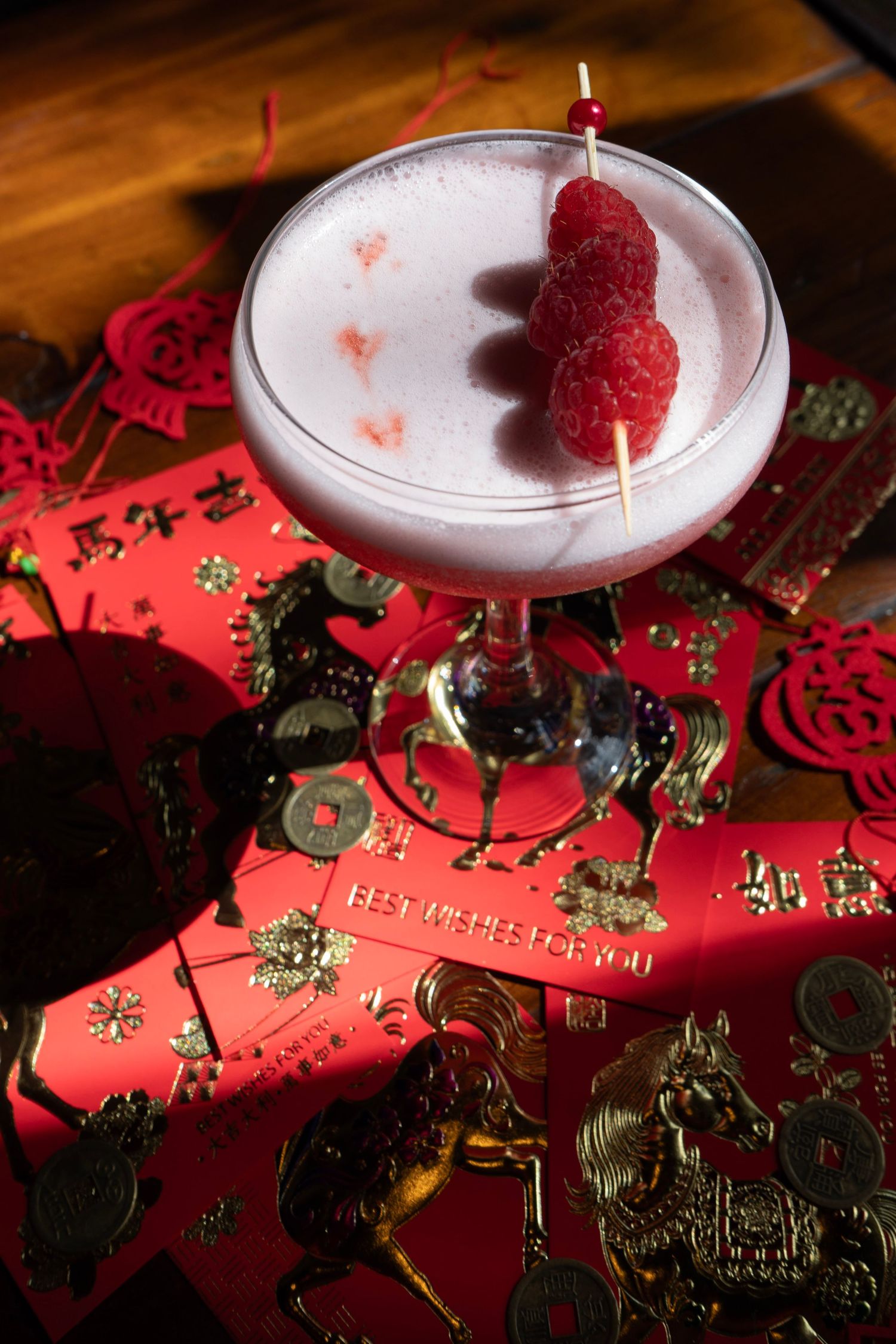 Frothy pink cocktail garnished with fresh raspberries on a skewer surrounded by Chinese New Year decor.