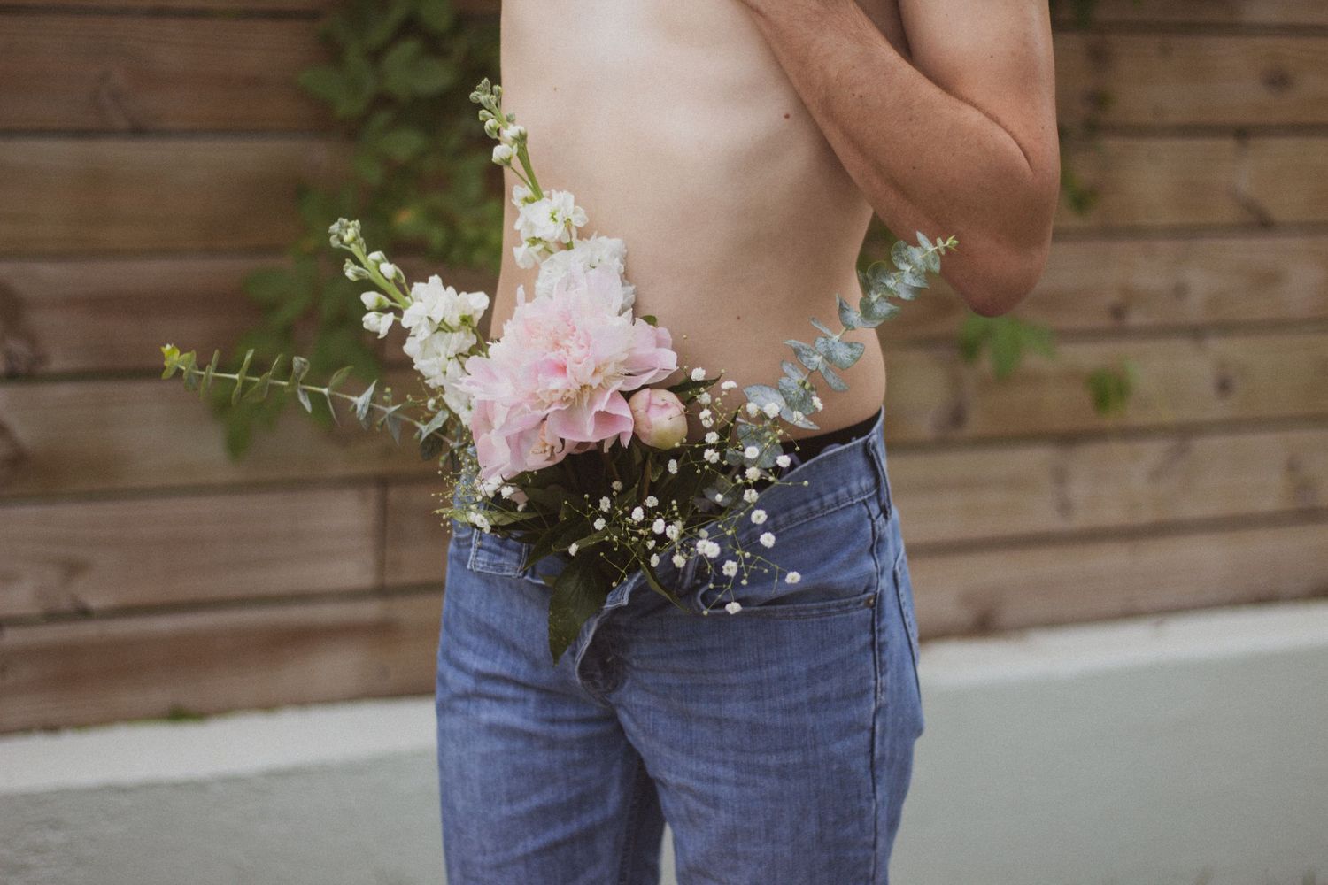 A shirtless person holds a bouquet of pink and white flowers at their waist while wearing blue jeans outdoors.