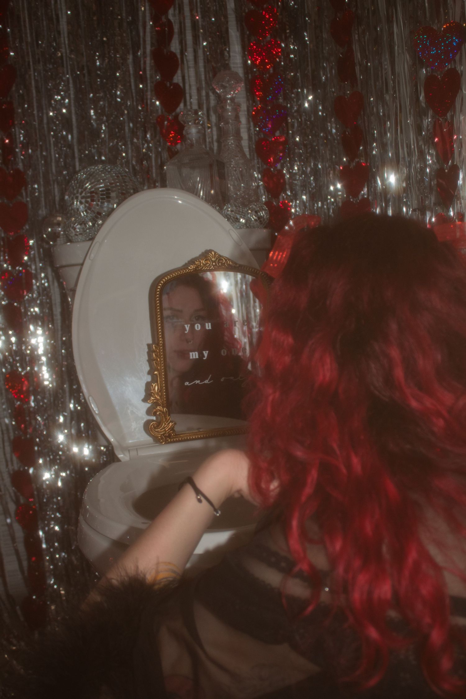 Red-haired woman looking into ornate mirror surrounded by silver tinsel and red heart decorations in moody setting.