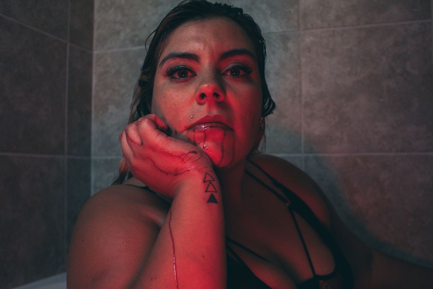A woman in black lingerie sits in a bathtub under red lighting, resting her chin on her tattooed hand.