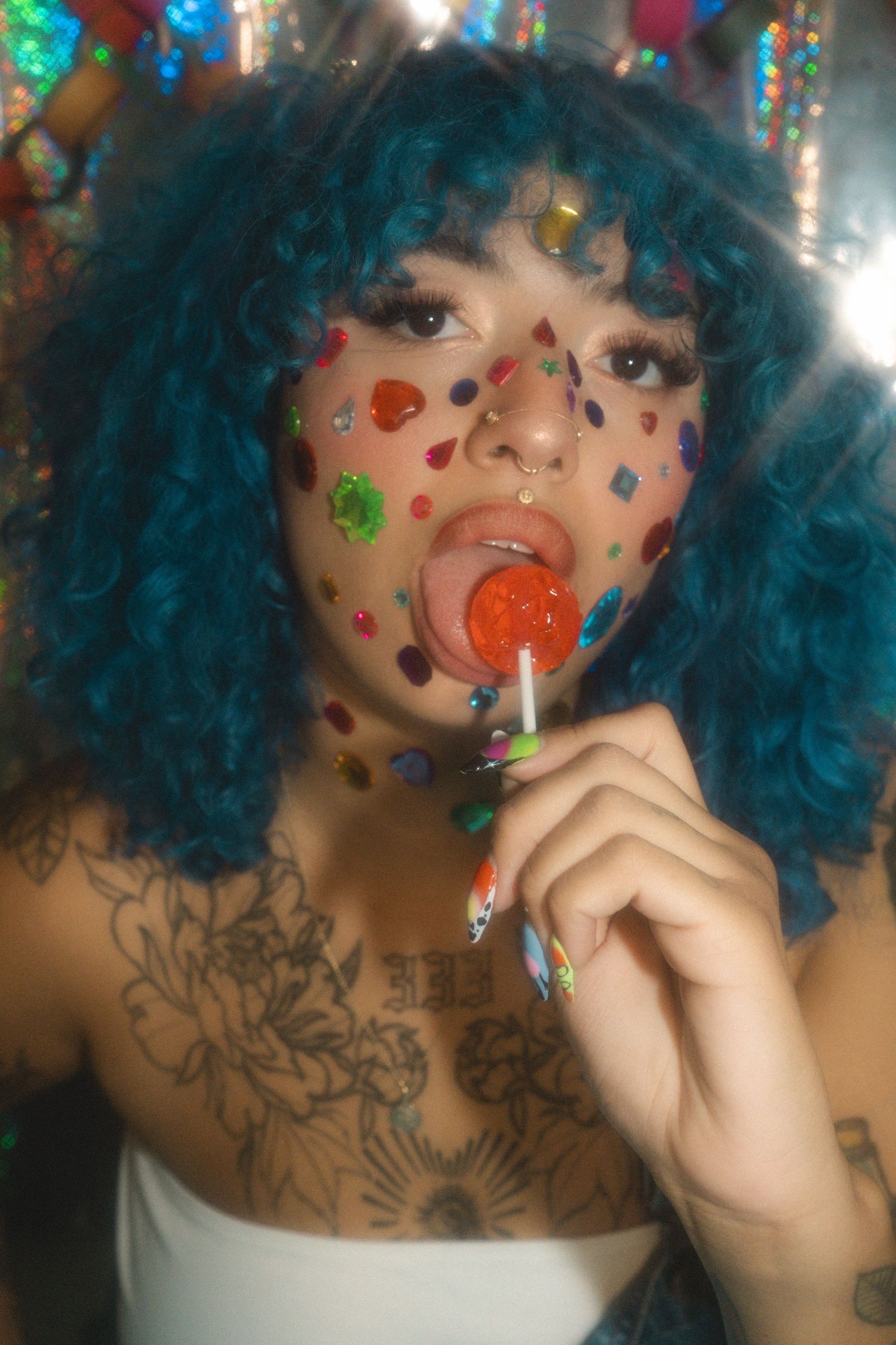 Woman with blue curly hair, colorful face gems, tattoos, licking a red lollipop in a vibrant, glittery setting.