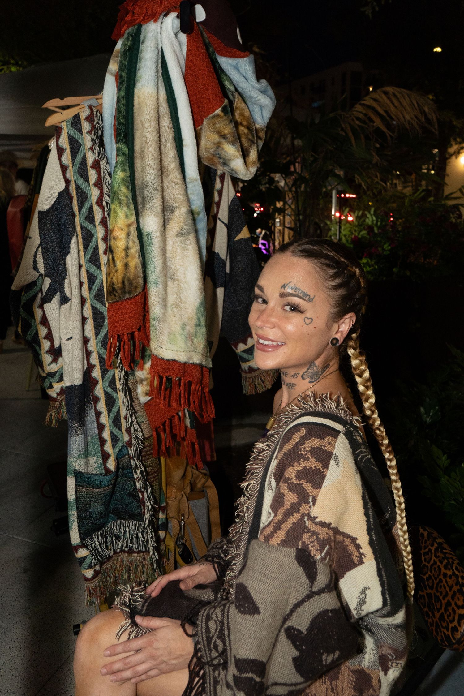 Woman with braided hair and face gems poses in patterned wrap beside colorful fringed textiles display.