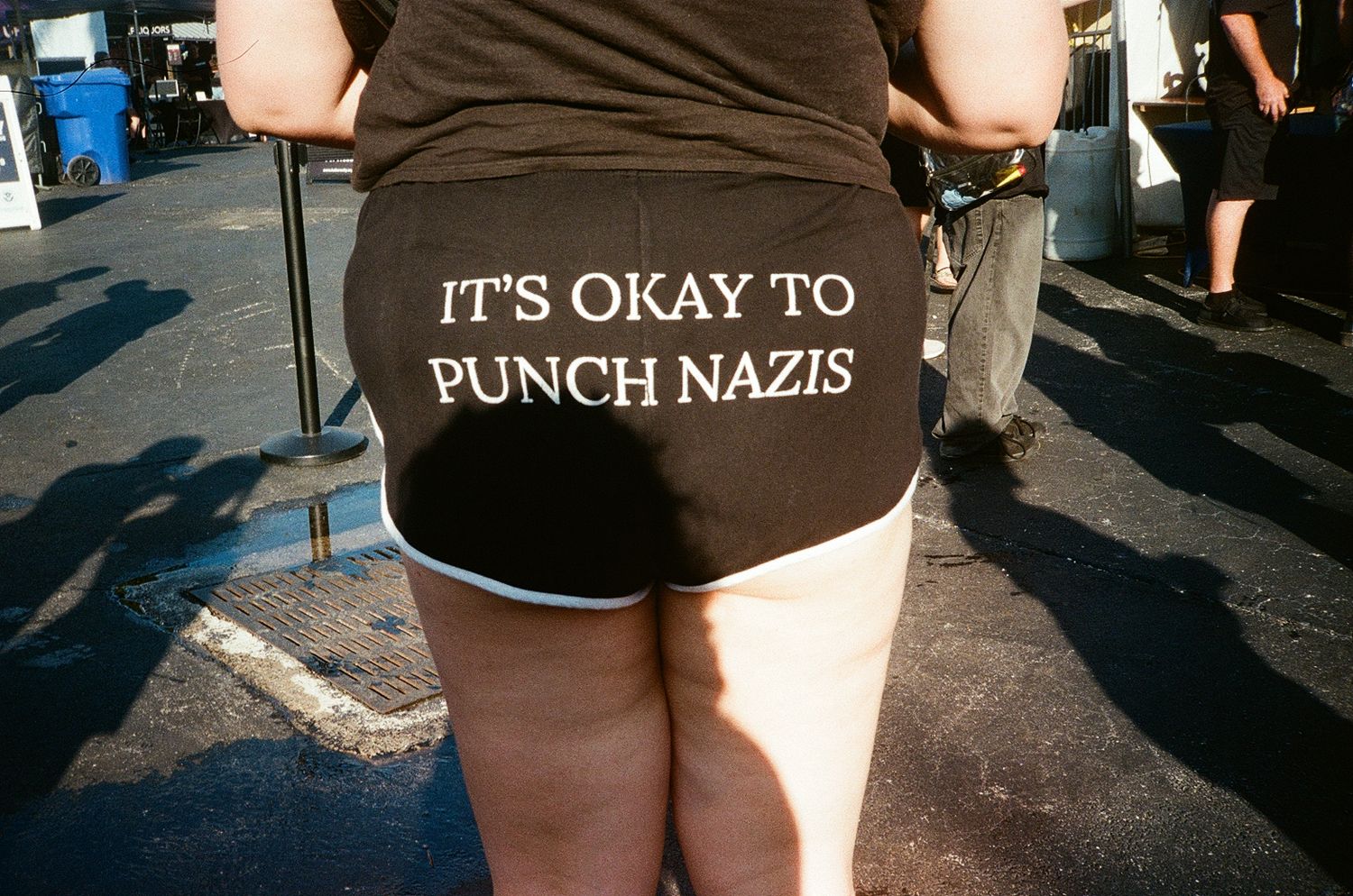 Person wearing black shorts with white text reading IT'S OKAY TO PUNCH NAZIS at an outdoor event.