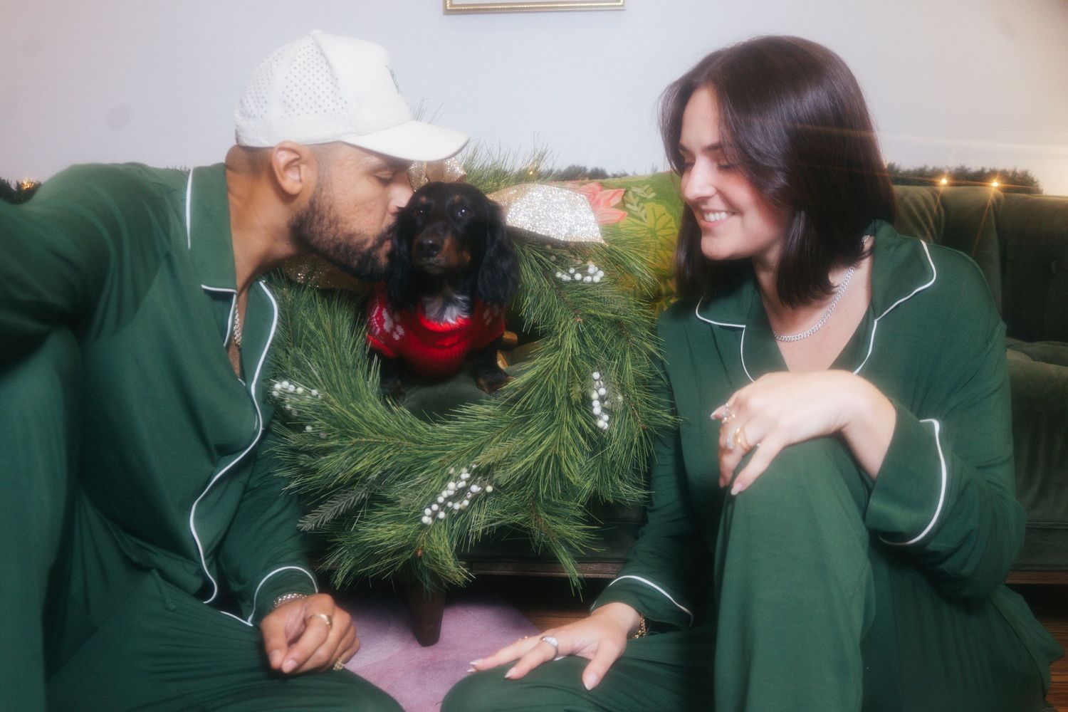 Couple in green pajamas kissing their dachshund wrapped in a festive pine wreath.