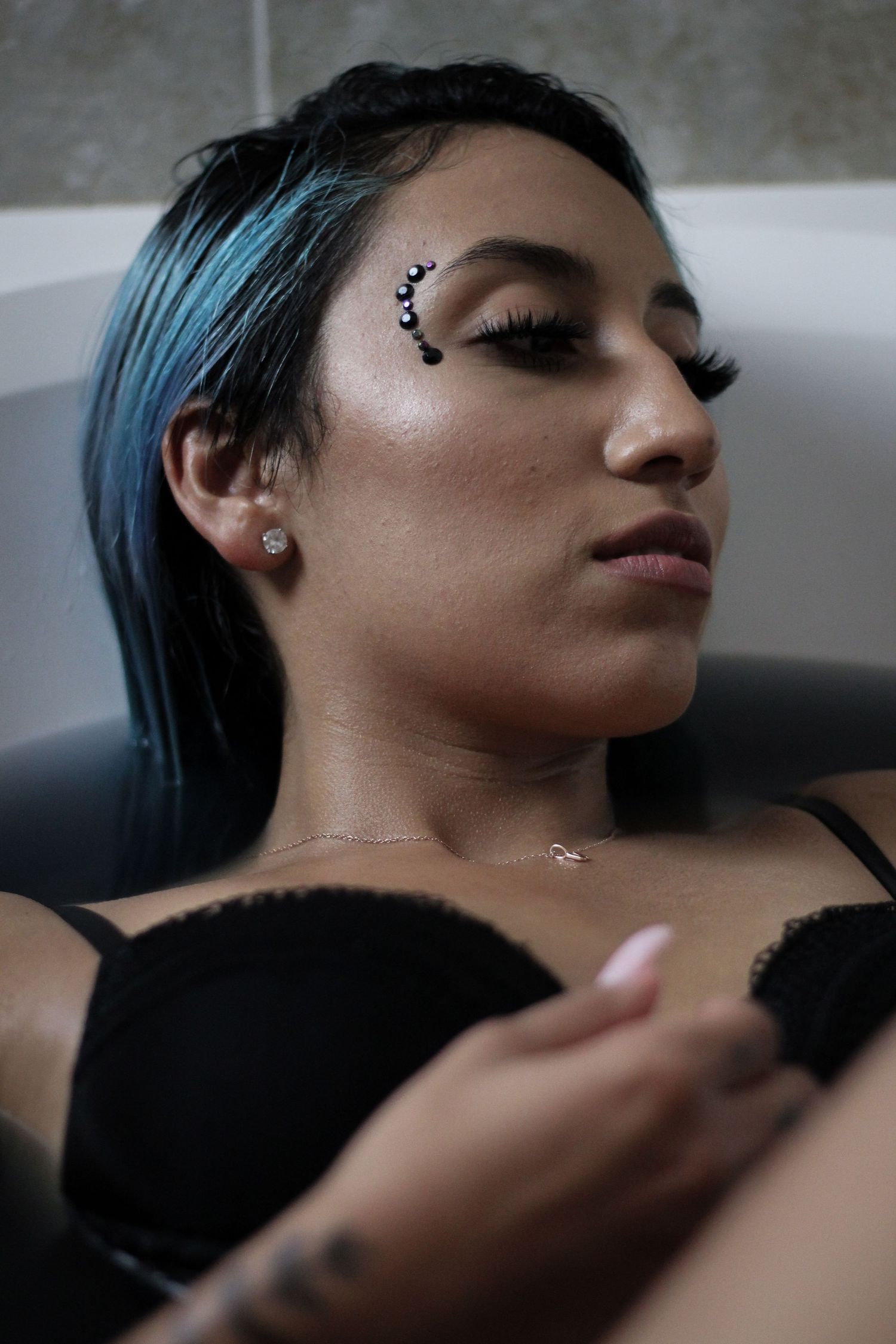 Woman with blue hair and face gems reclines in bathtub wearing black lingerie.