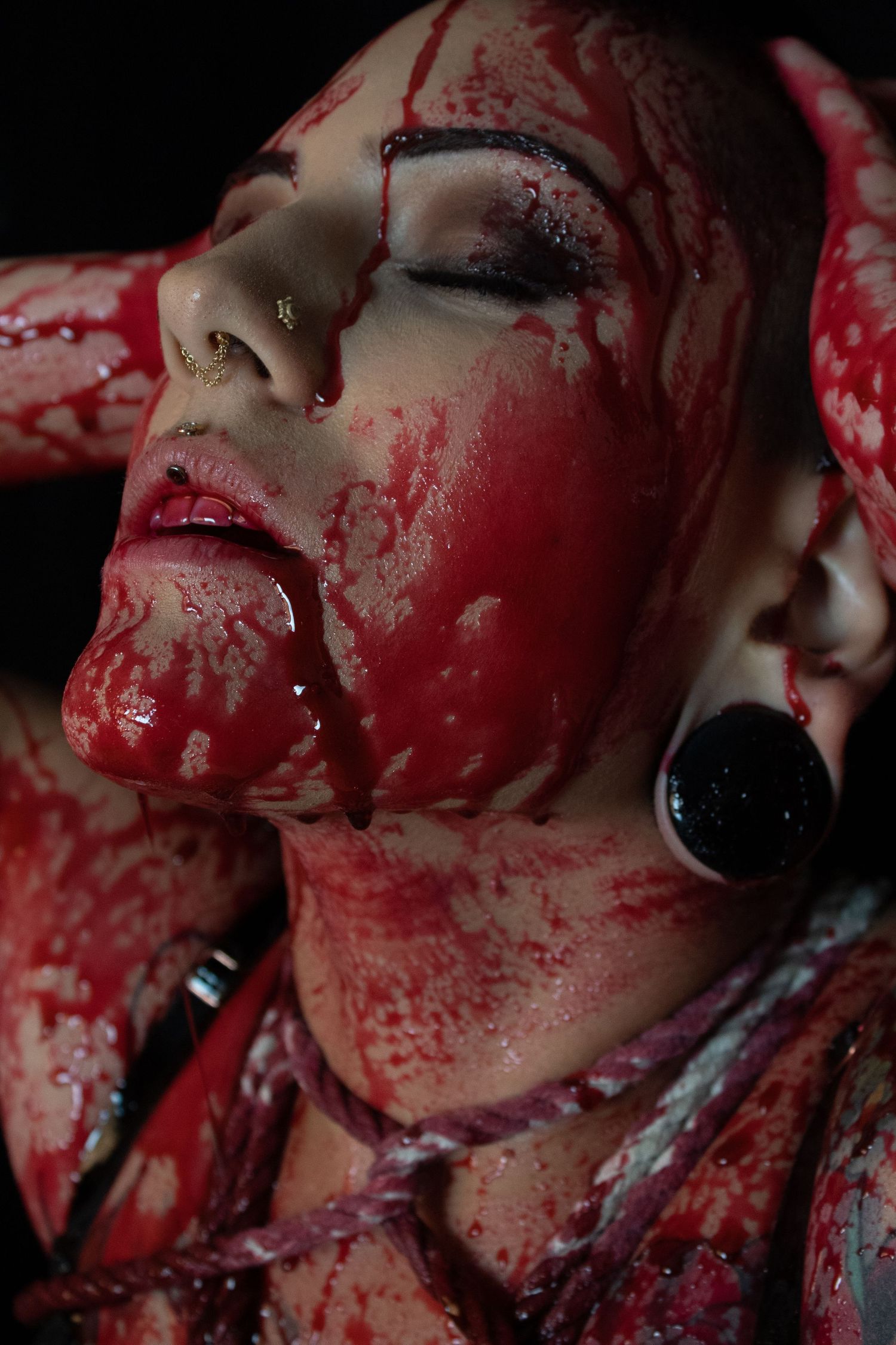 Close-up of a woman covered in red paint simulating blood, with gauge earrings, in dramatic horror-themed artistic photography.