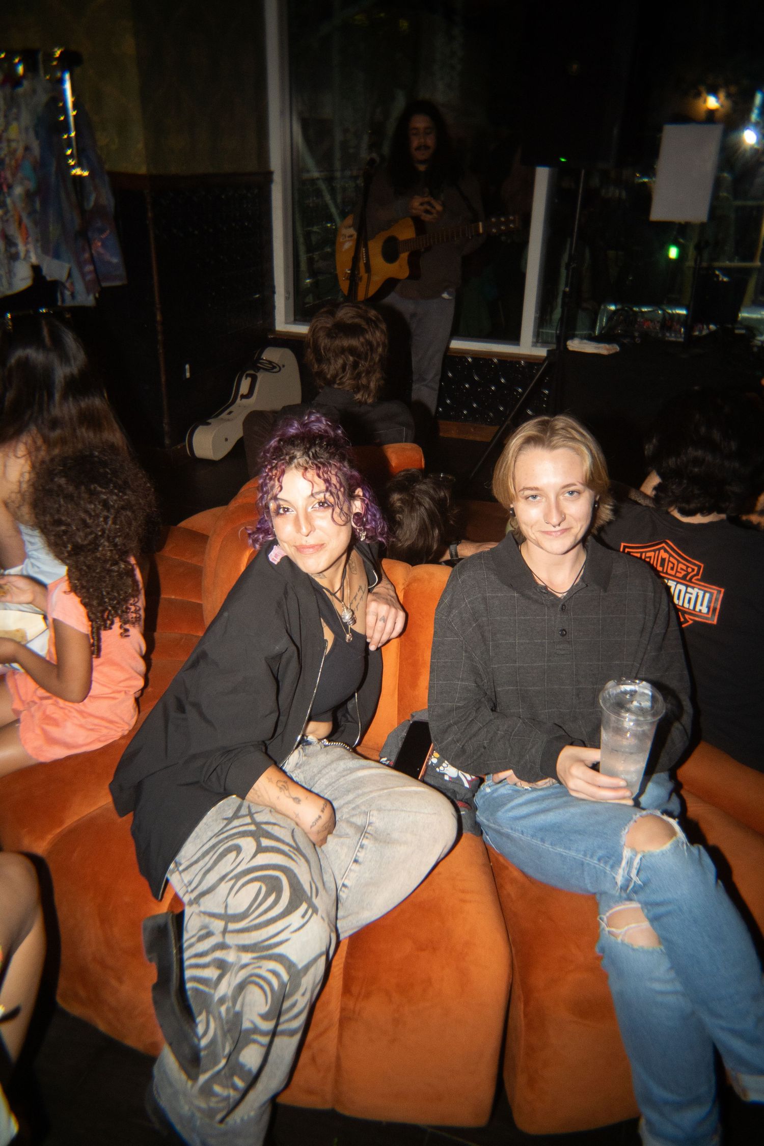 Two young women sitting on an orange couch at a live music event, with a guitarist performing in the background.