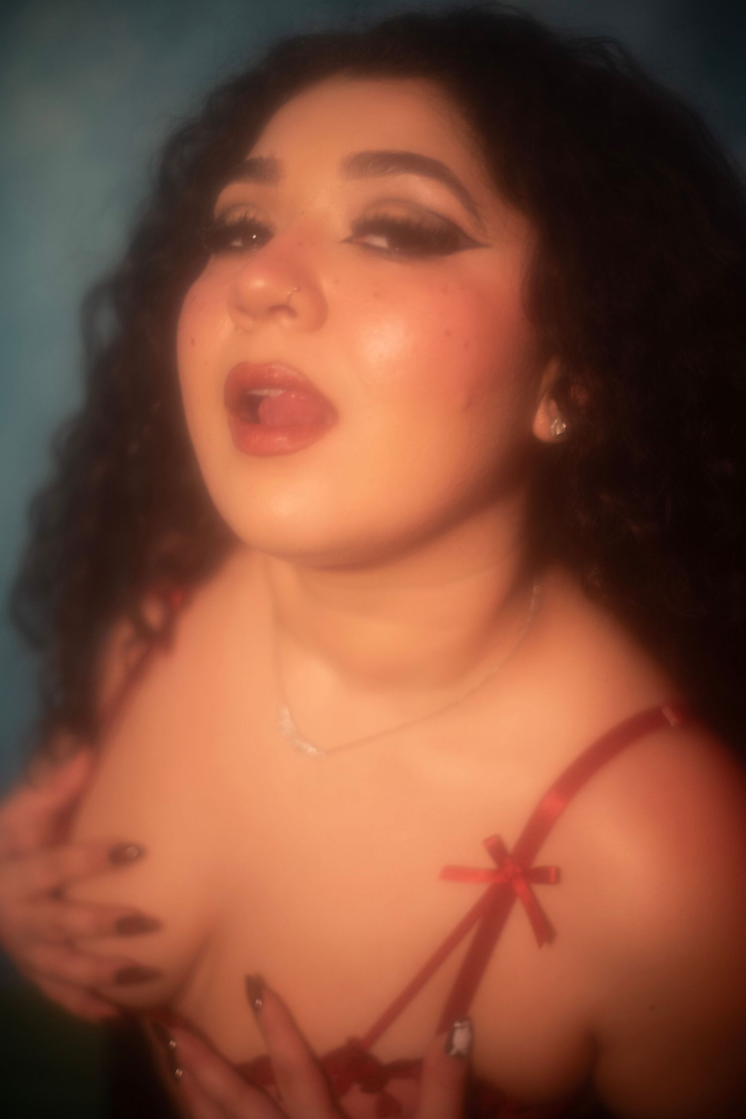 Curly-haired woman in red lingerie with dramatic makeup posing expressively under warm red lighting.