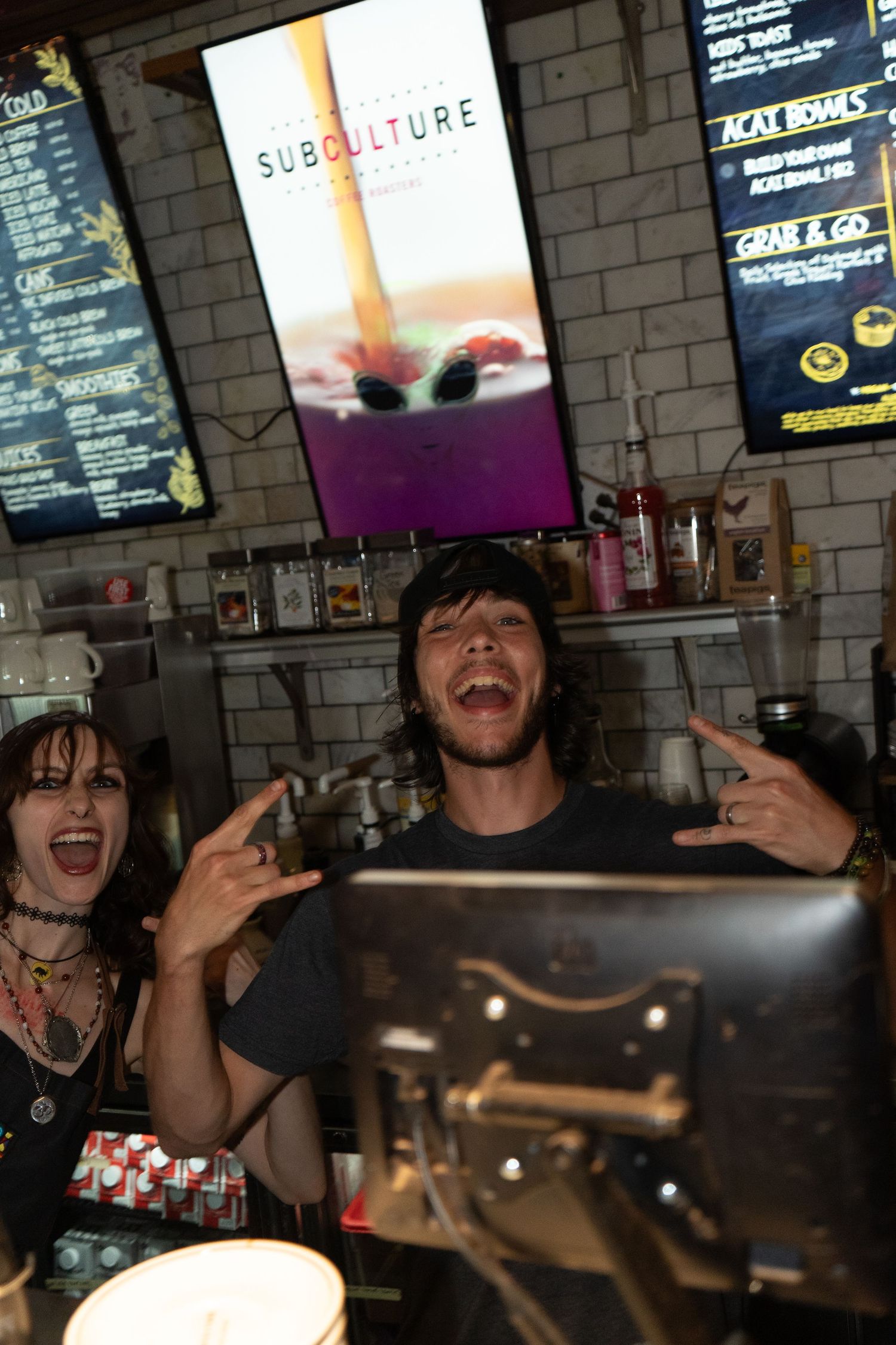 Two rock fans flash devil horns gestures at Subculture cafe, posing energetically behind the counter.