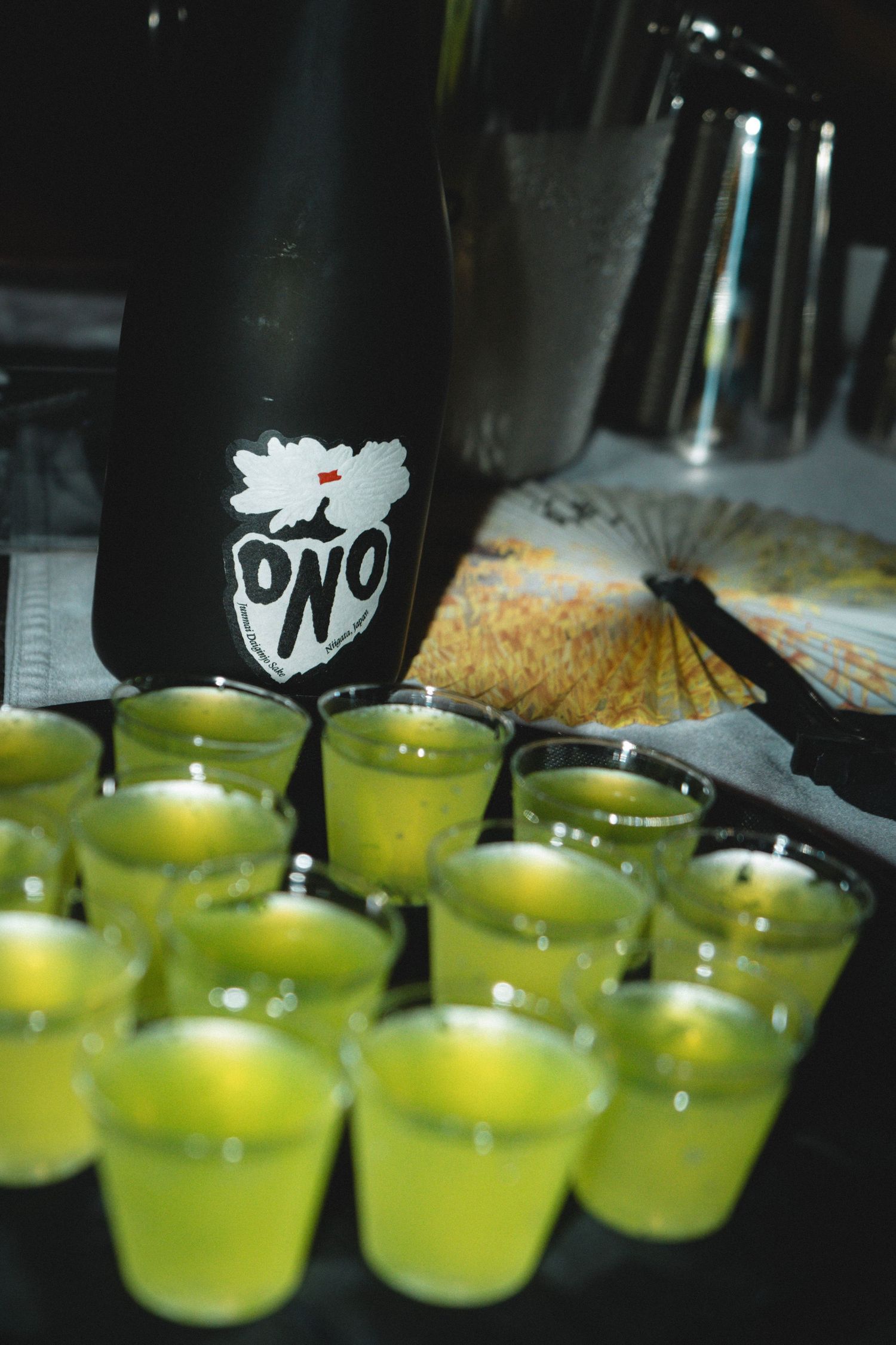 Green sake shots lined up in front of a black ONO sake bottle at a bar event.