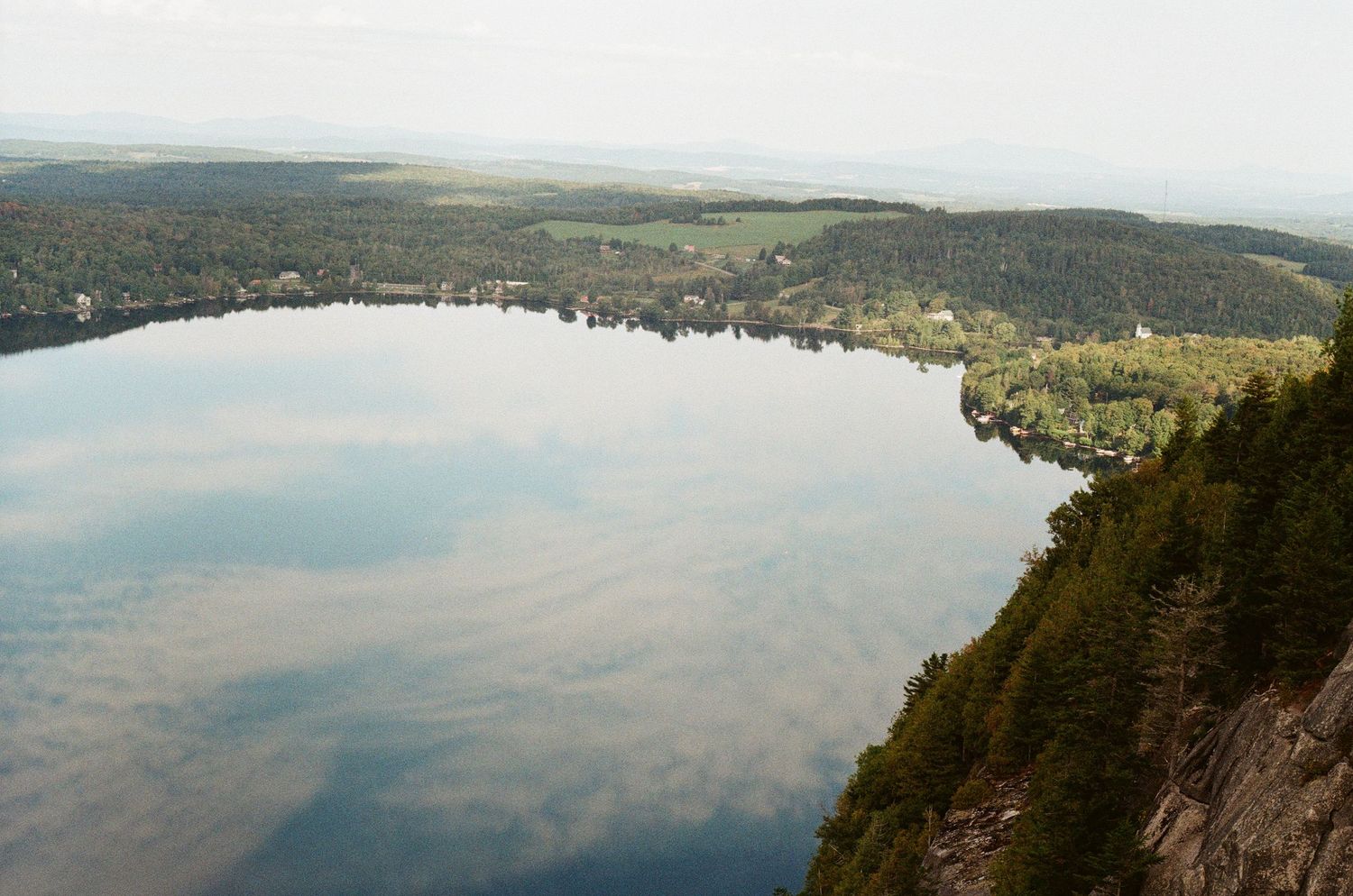 Aerial panoramic view of a large calm lake reflecting cloudy skies, surrounded by dense forest and distant hills.