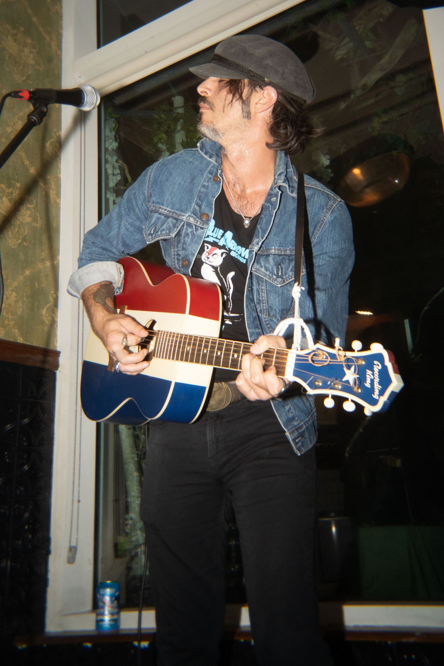 Male musician performing with a red, white, and blue guitar while wearing a denim jacket and black cap indoors.