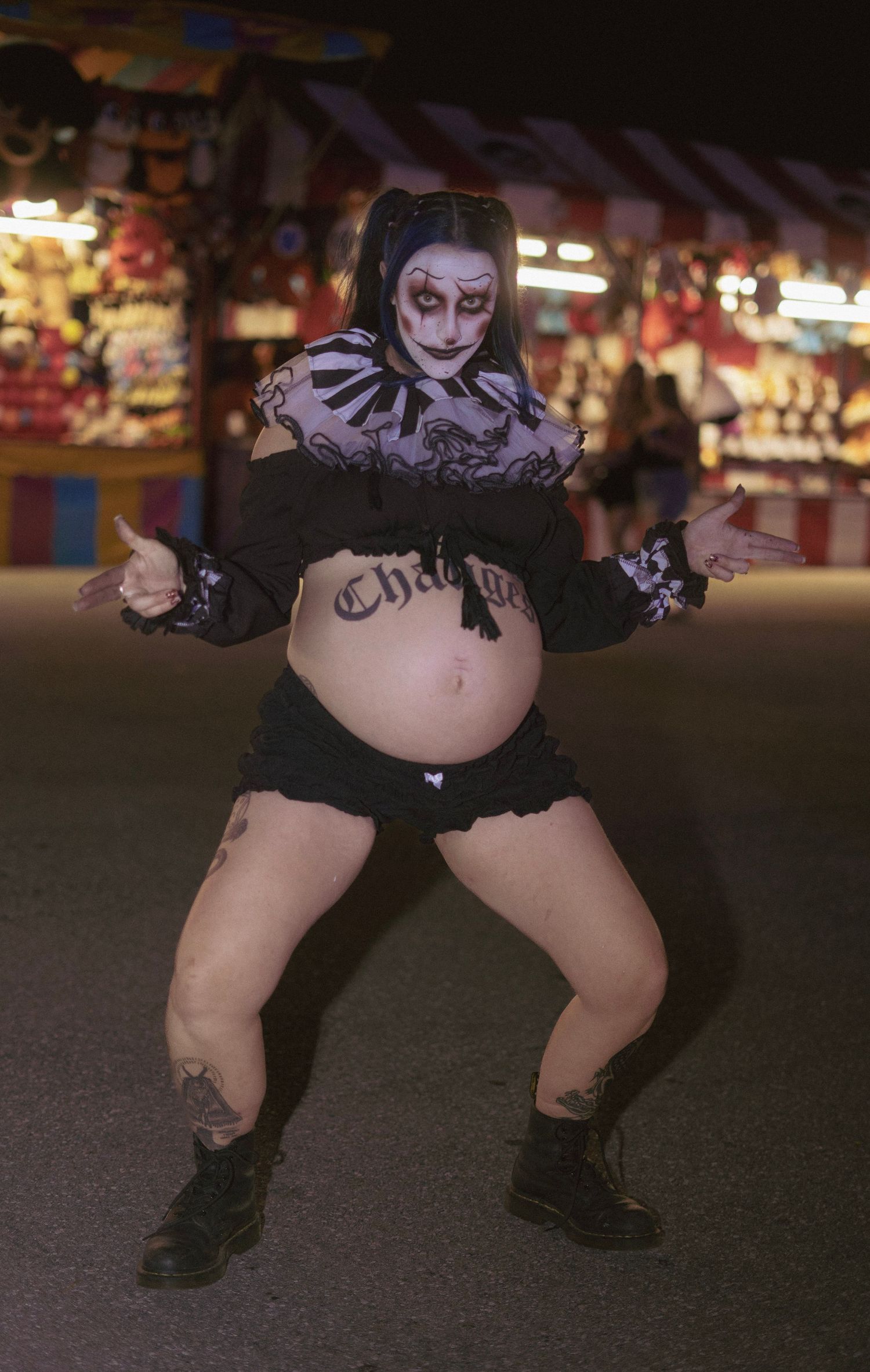 Pregnant woman in clown face paint with blue pigtails and black shorts poses at an outdoor carnival at night.