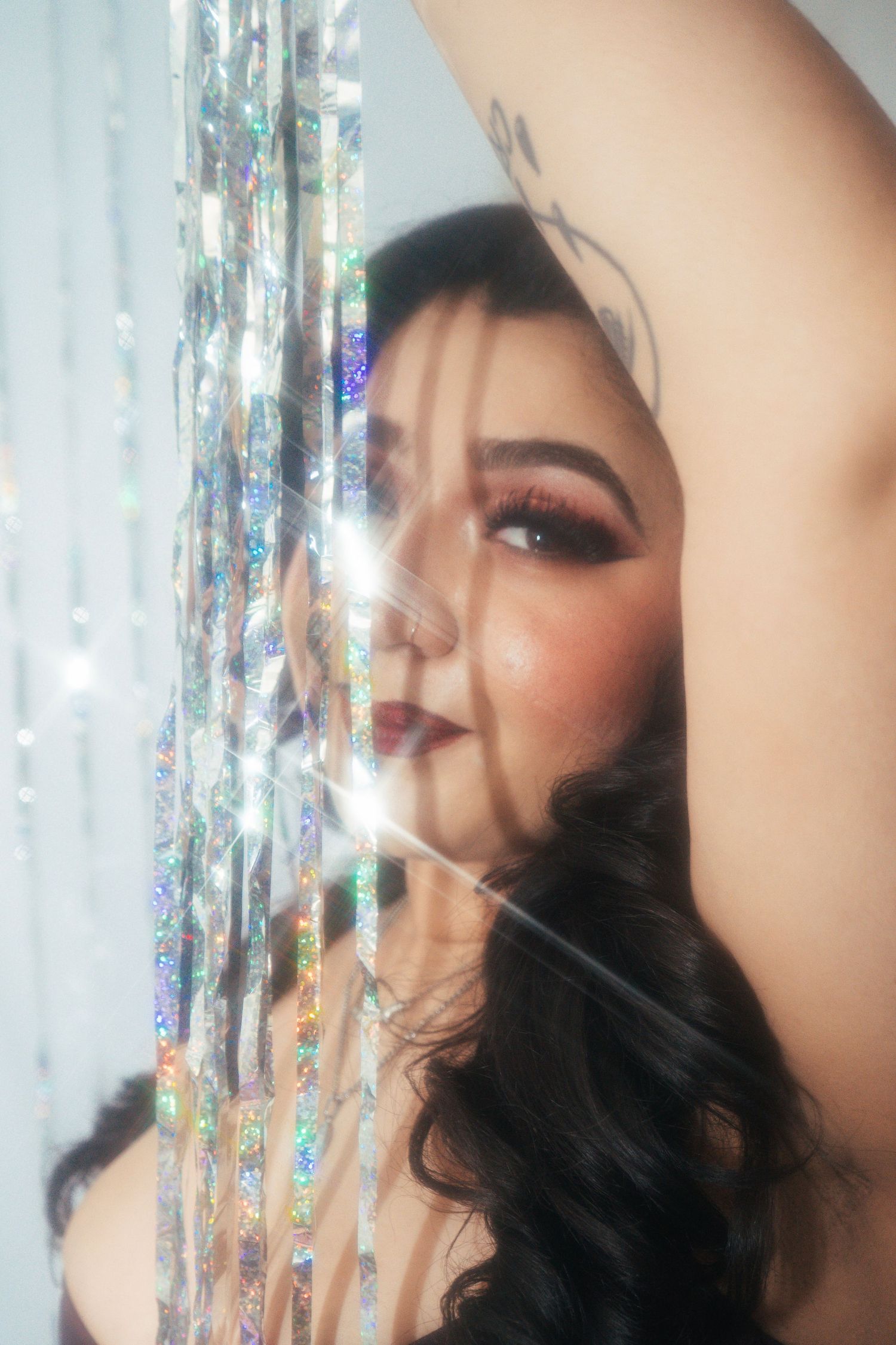 Woman with bold makeup posing behind sparkling holographic tinsel curtain with arm raised.