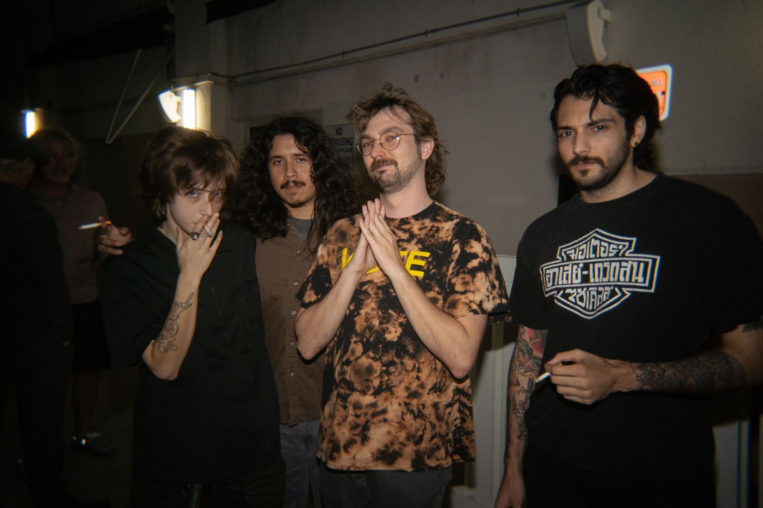Four young men with long hair pose together indoors, wearing casual band t-shirts in a dimly lit venue.