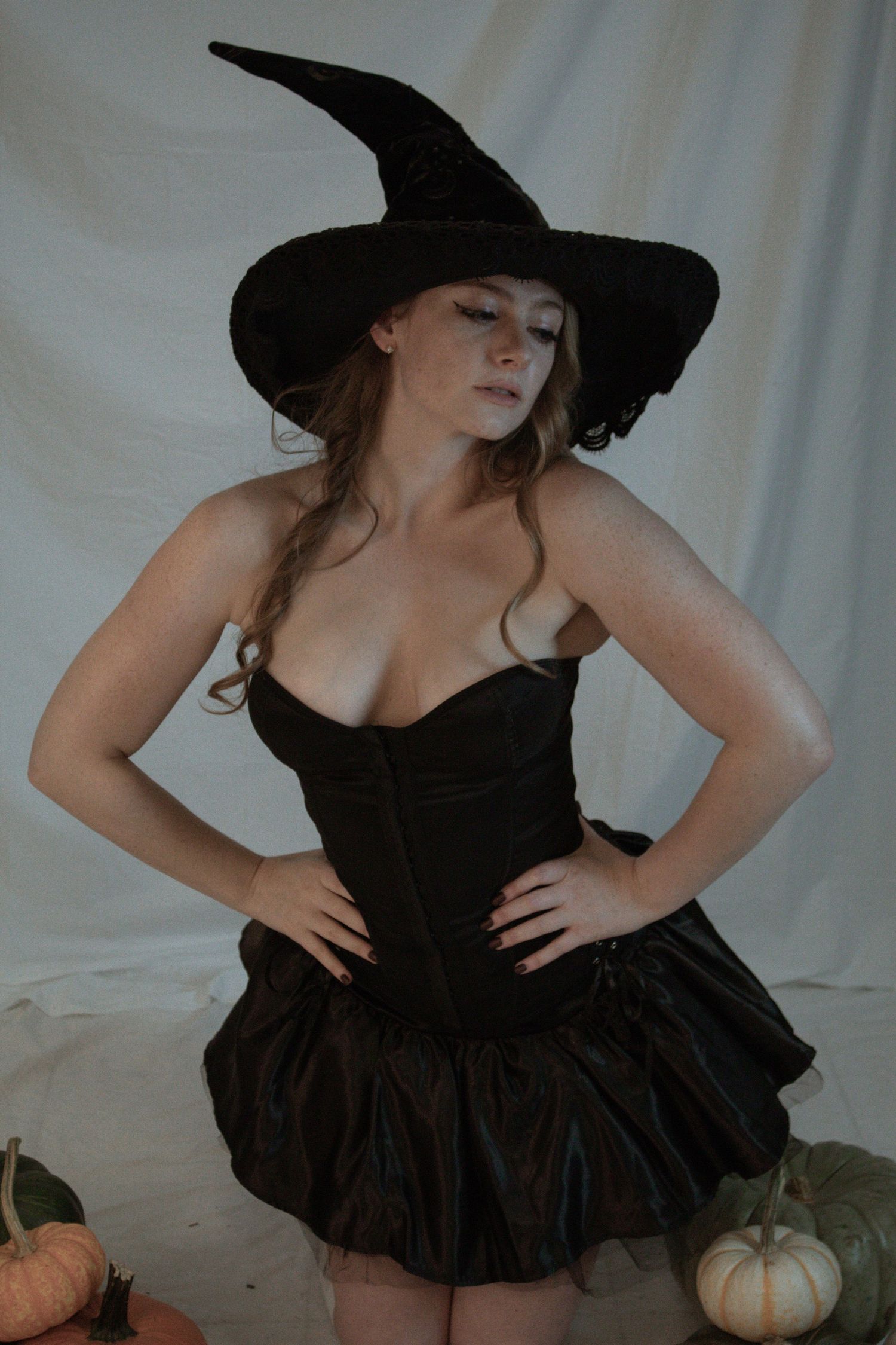 Woman in black witch costume with corset and mini skirt posing with pumpkins in a Halloween photoshoot.