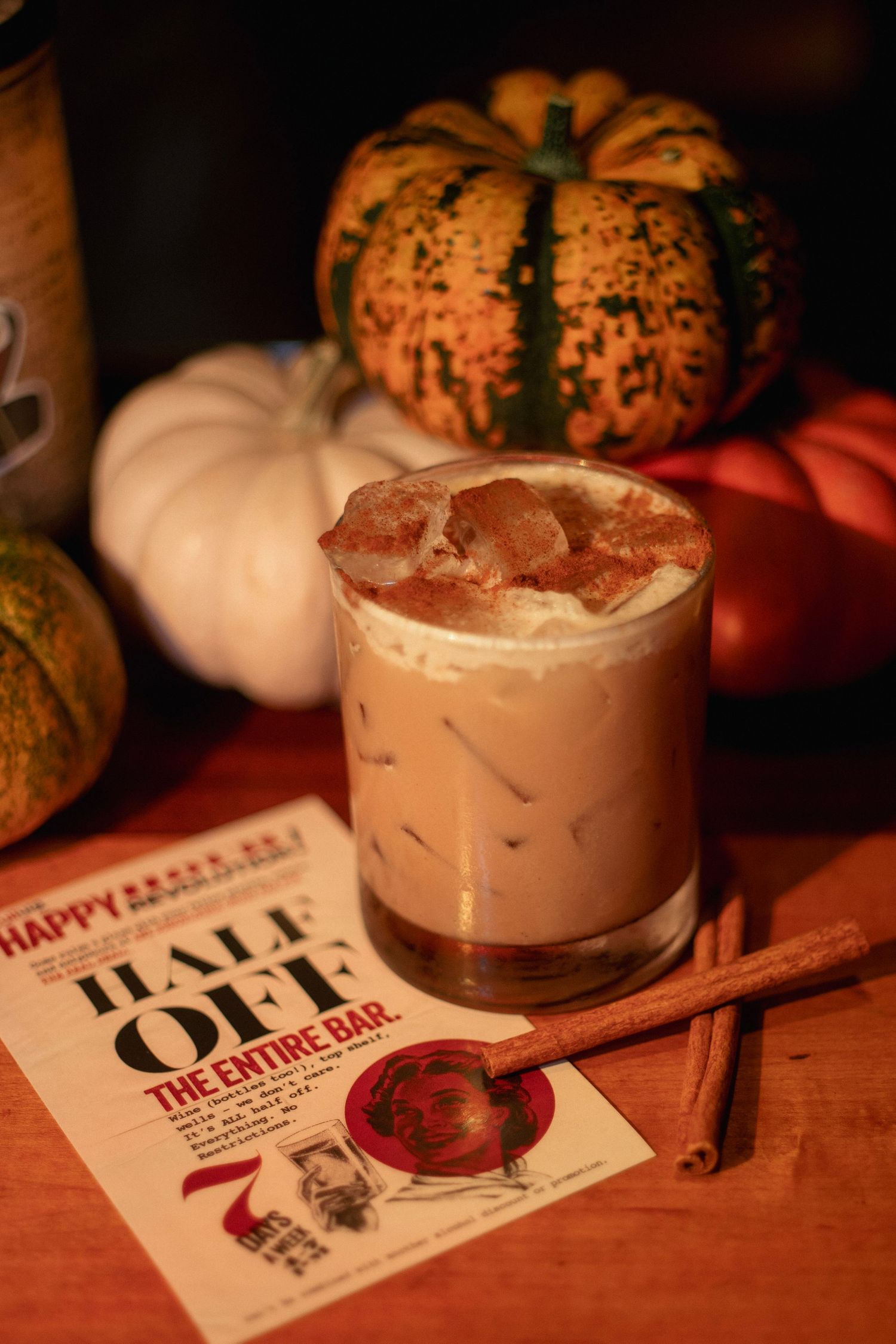 Creamy iced cocktail garnished with cinnamon sticks beside a half-off bar promotion flyer and decorative pumpkins.