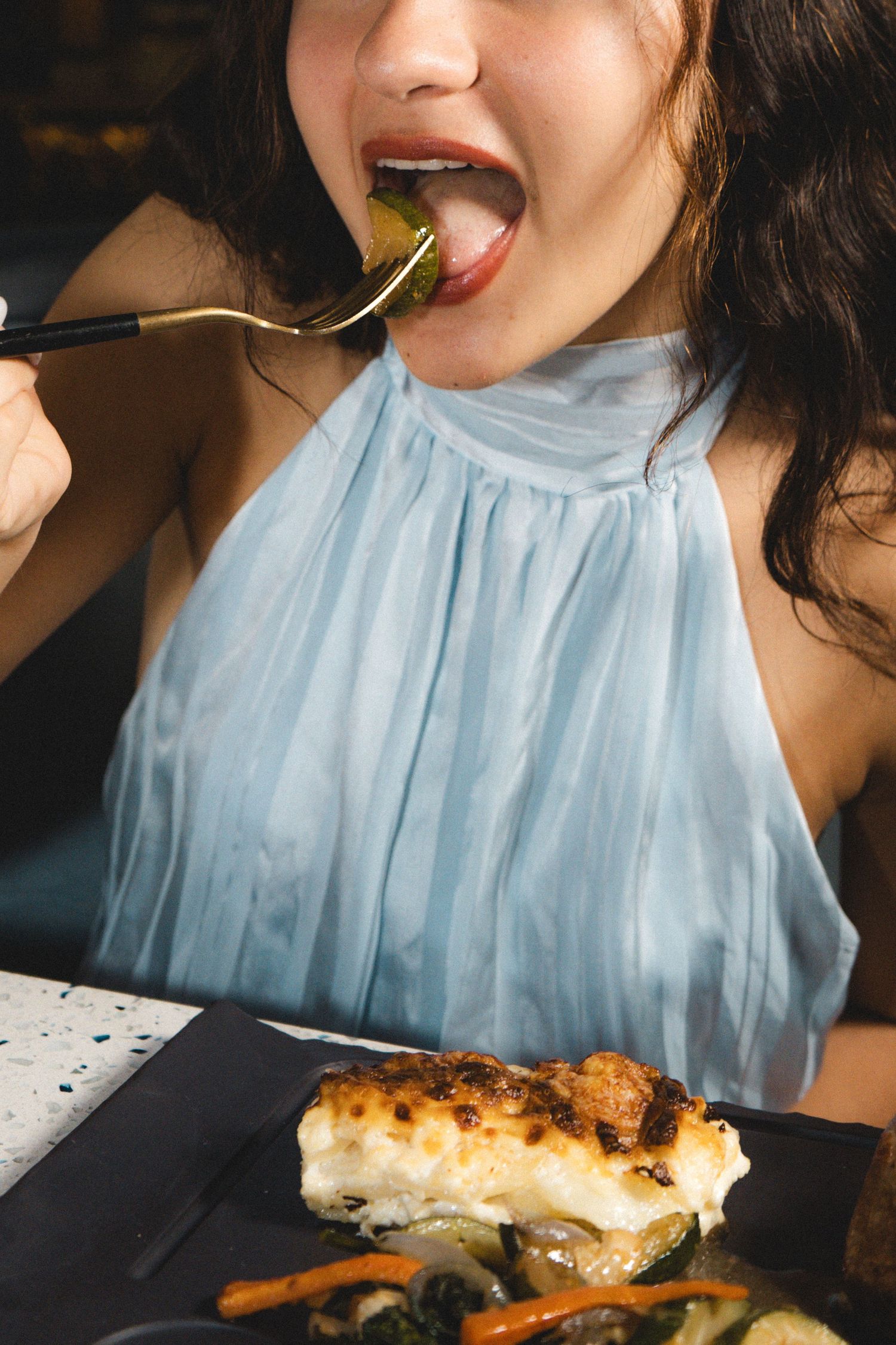 Woman in blue halter top eating fish dish with fork at restaurant table with roasted vegetables.