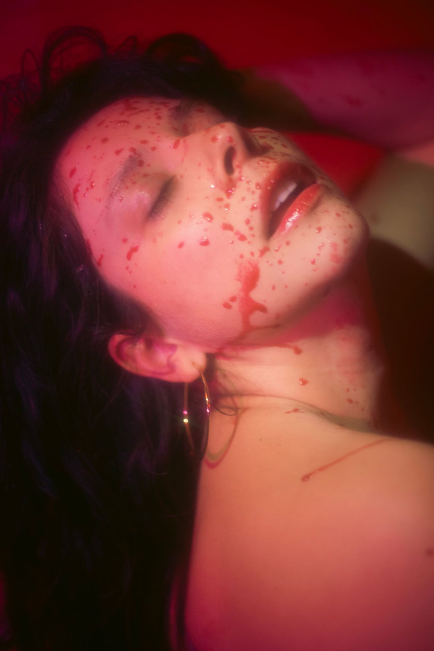 A woman with closed eyes and simulated blood splatter on her face lies back in a horror-themed shoot.